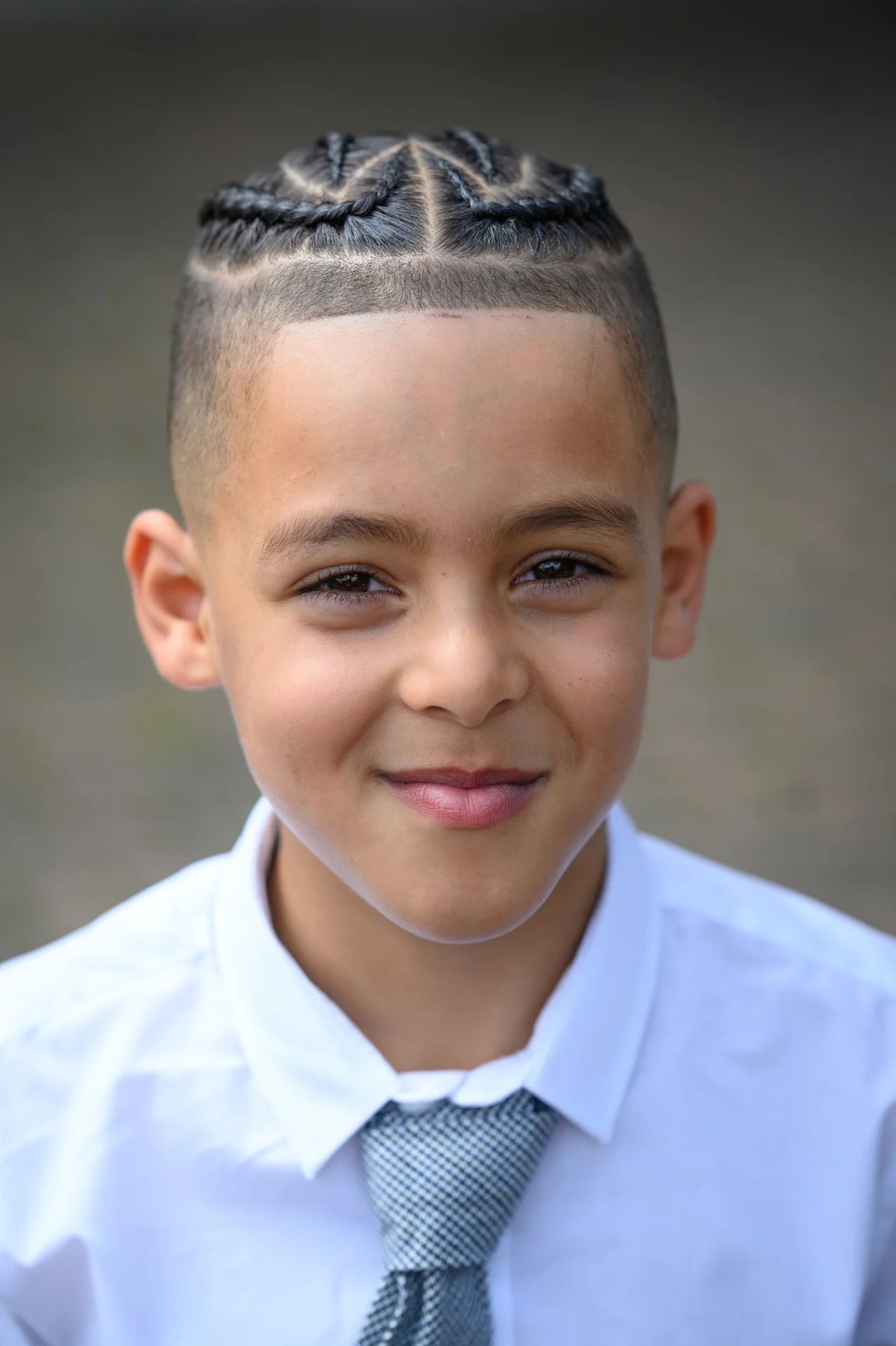 A young page boy with neat braided hair and a patterned tie smiles gently for the camera.