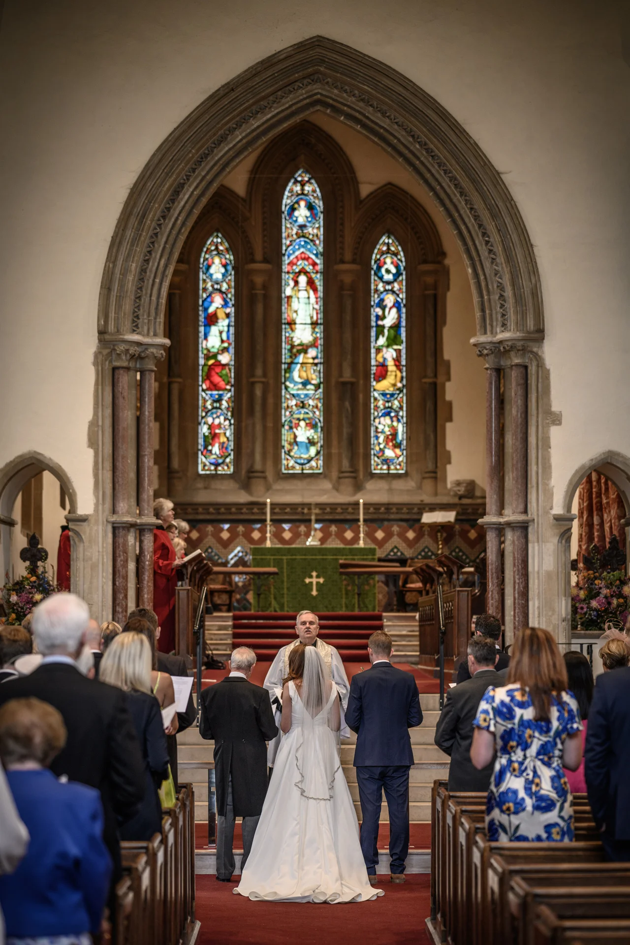 A back-view shot of a bride and groom standing before the altar during a traditional church wedding ceremony beneath a magnificent stone archway.