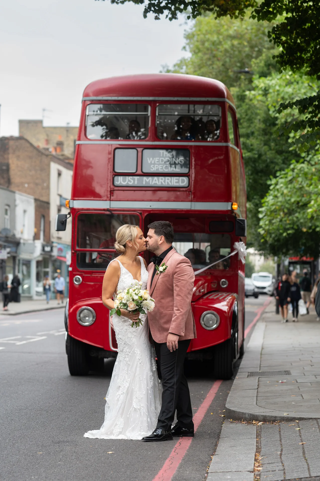 A newlywed couple shares a tender kiss on a London street in front of a classic red double decker bus displaying a just married sign.