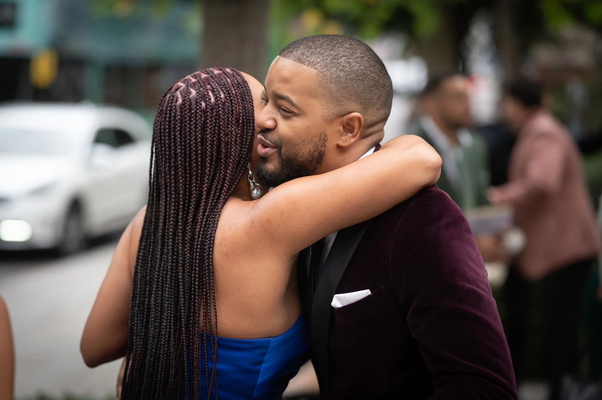 A groom wearing a burgundy velvet tuxedo embraces a guest with braided hair in a warm, tender moment during an outdoor city wedding celebration.