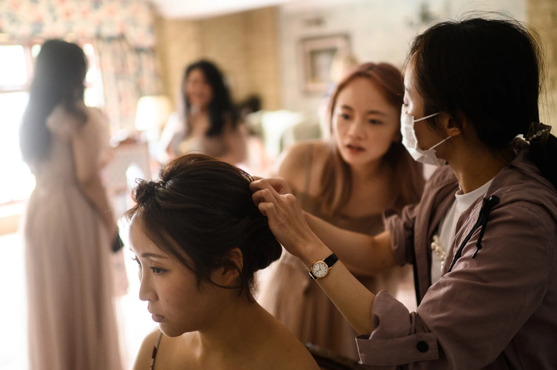 A bride has her hair styled by a professional while bridesmaids gather in a brightly lit room during morning wedding preparations.