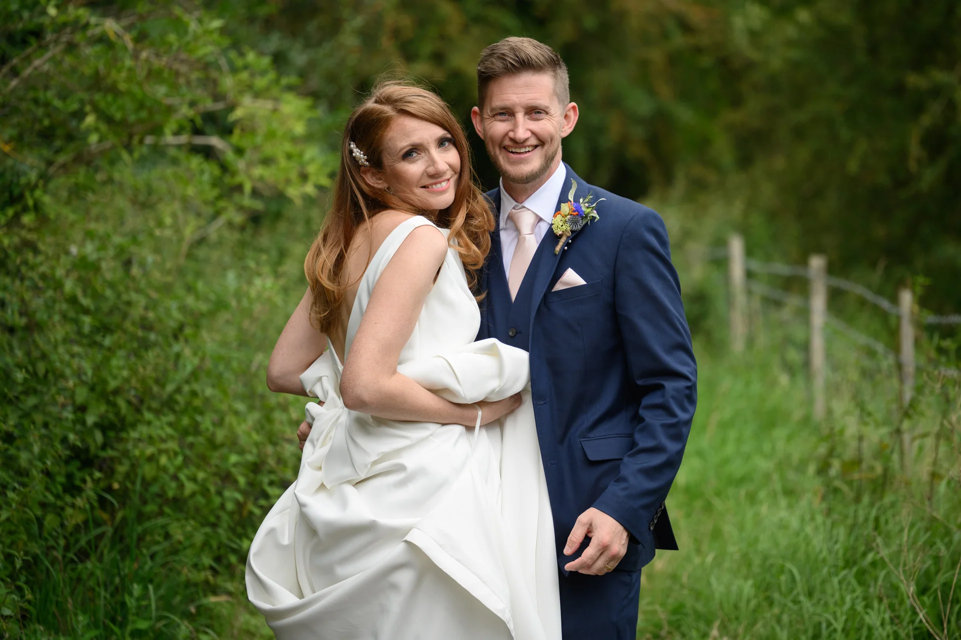 A newlywed couple smiles warmly together outdoors in a lush green woodland setting, with the bride wearing a sleek satin gown and the groom in a classic navy suit.
