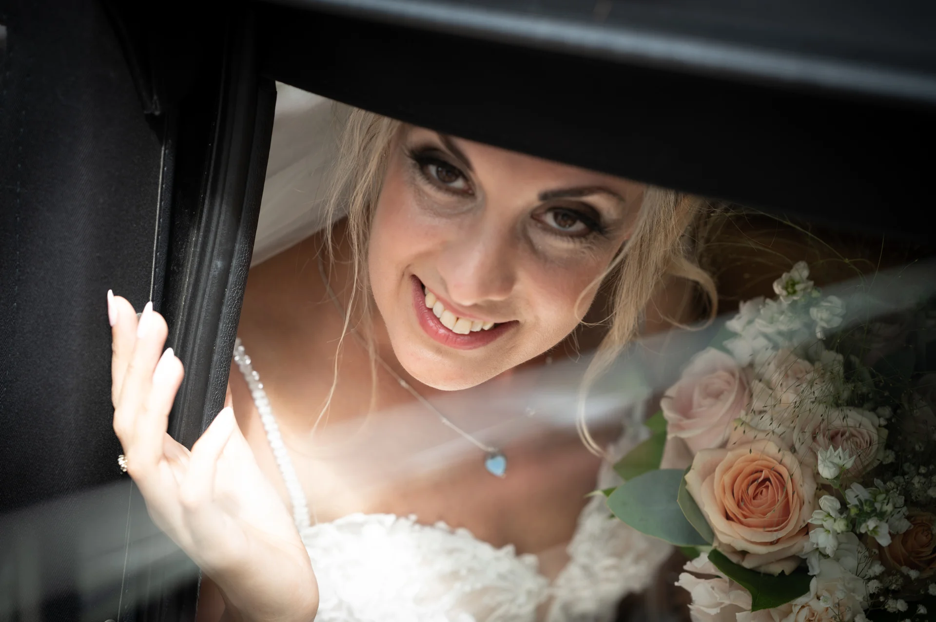 A smiling bride looks out through the window of a classic wedding car, holding a soft floral bouquet of roses and eucalyptus.