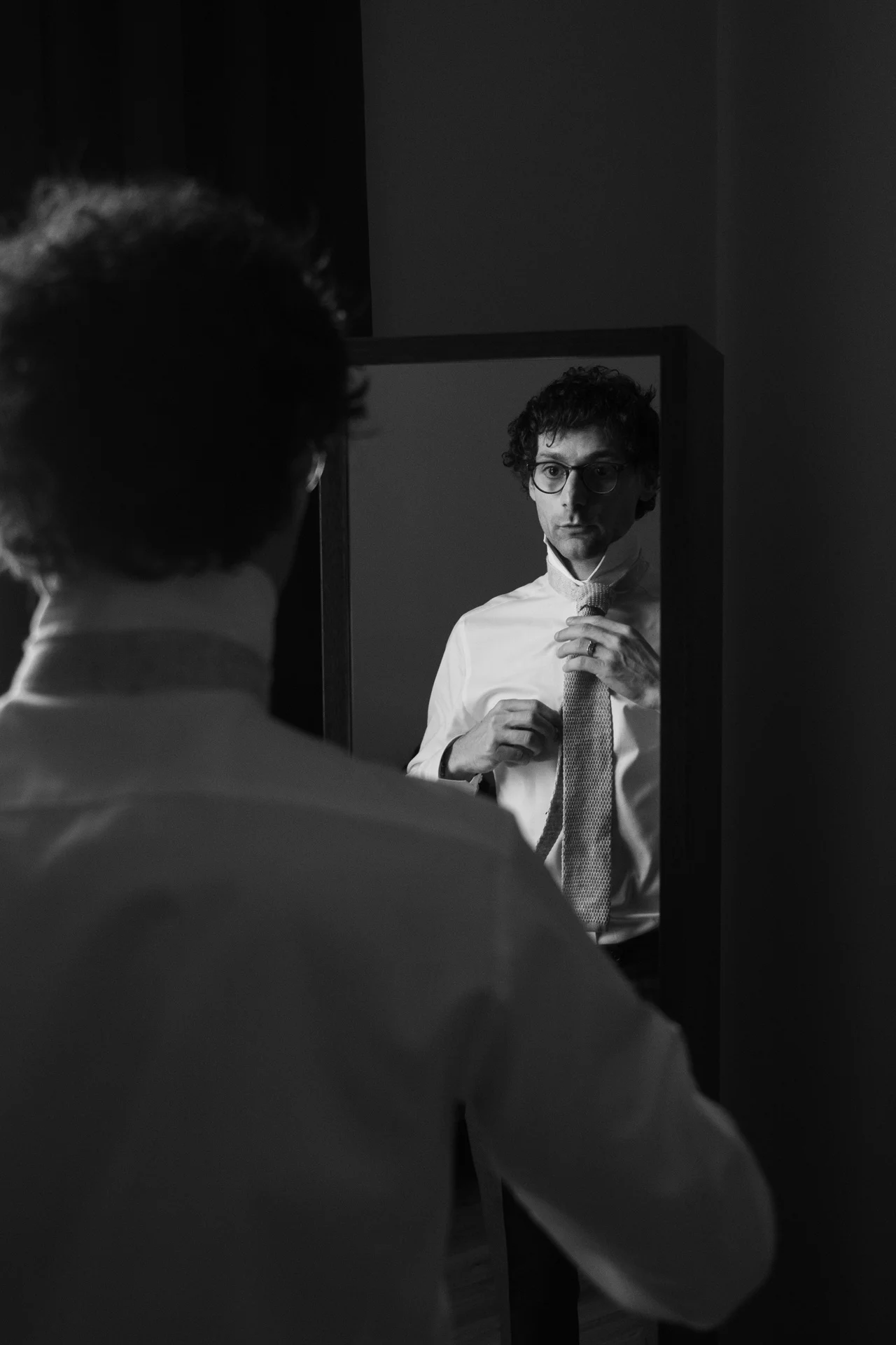 A groom stands in front of a mirror adjusting his knitted tie during morning wedding preparations.