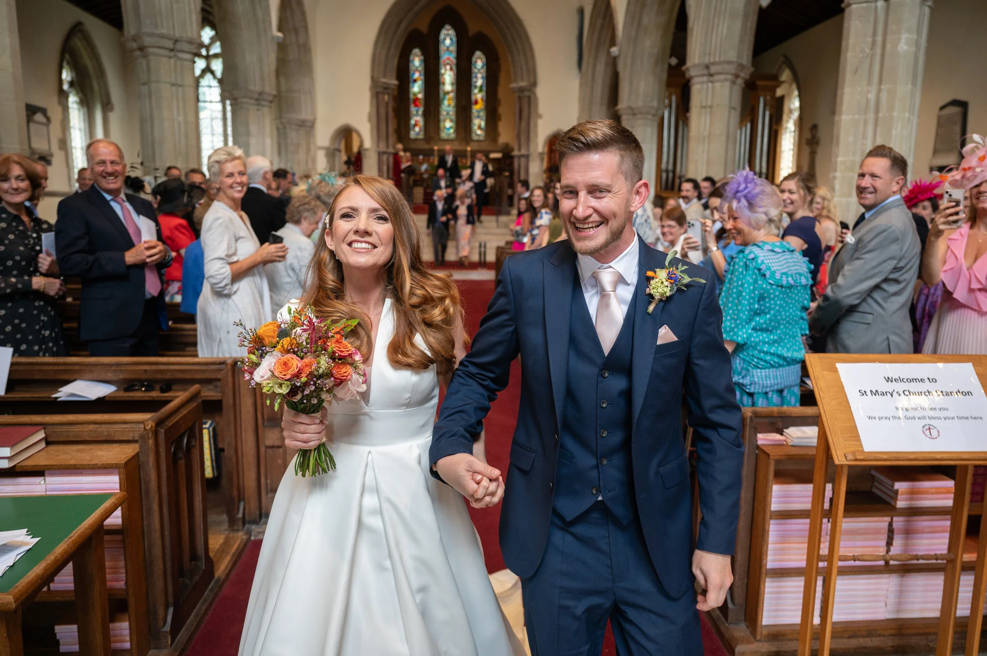 A smiling newlywed couple walks hand in hand down the aisle of a traditional church after their wedding ceremony, surrounded by cheering guests.