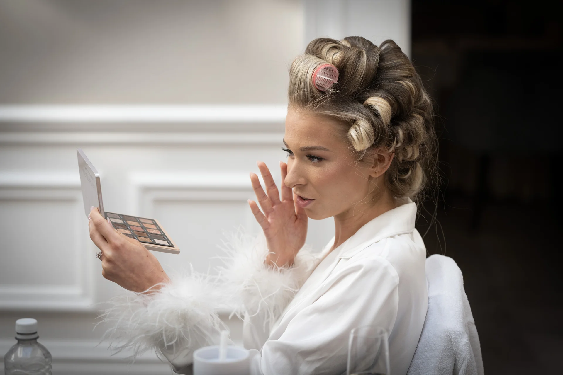 A bride with hair in rollers and wearing a white robe with feathered cuffs checks her makeup in a small compact mirror during morning preparations.
