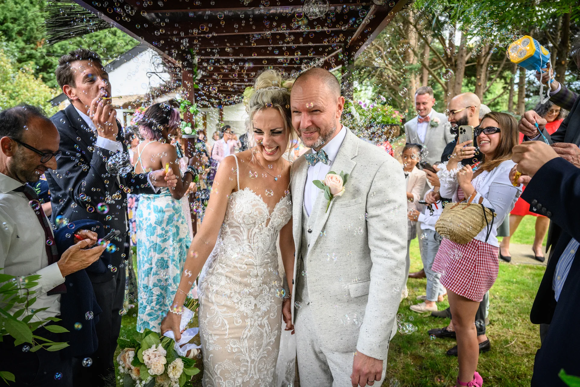 A newlywed couple walks through a pergola as cheering guests blow bubbles around them during a joyful outdoor summer wedding celebration.