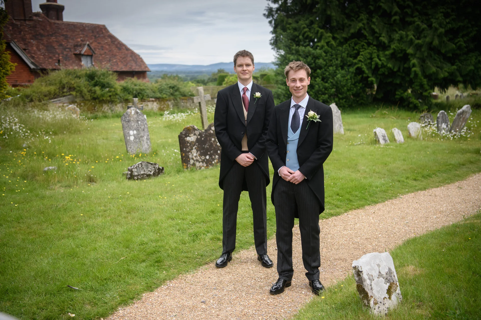 Two grooms dressed in traditional morning suits pose for a portrait along a gravel path in a historic churchyard.