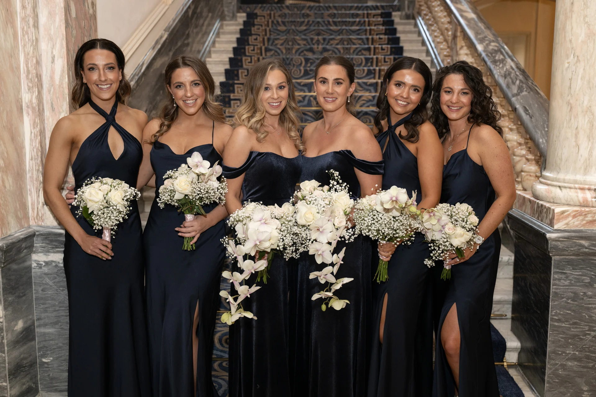 A group of six bridesmaids in matching navy blue dresses holding cascading bouquets of white roses and orchids stand on a grand indoor staircase.