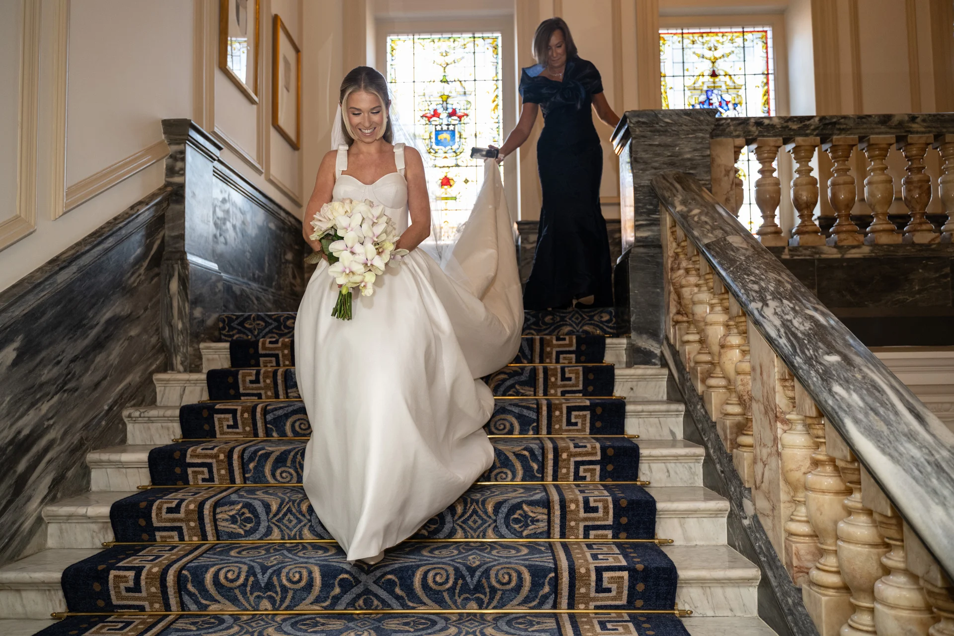 A smiling bride holding a bouquet of orchids descends a grand marble staircase with a patterned runner, while another woman follows closely behind carrying her dramatic train.