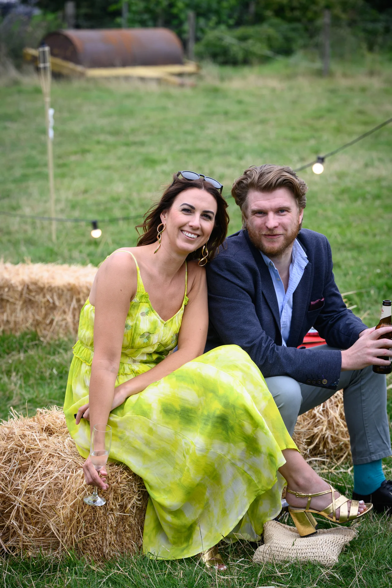 A well-dressed wedding guest in a vibrant yellow tie-dye dress and gold block heels sits on a hay bale with a man in a navy blazer outdoors, holding a glass of champagne.
