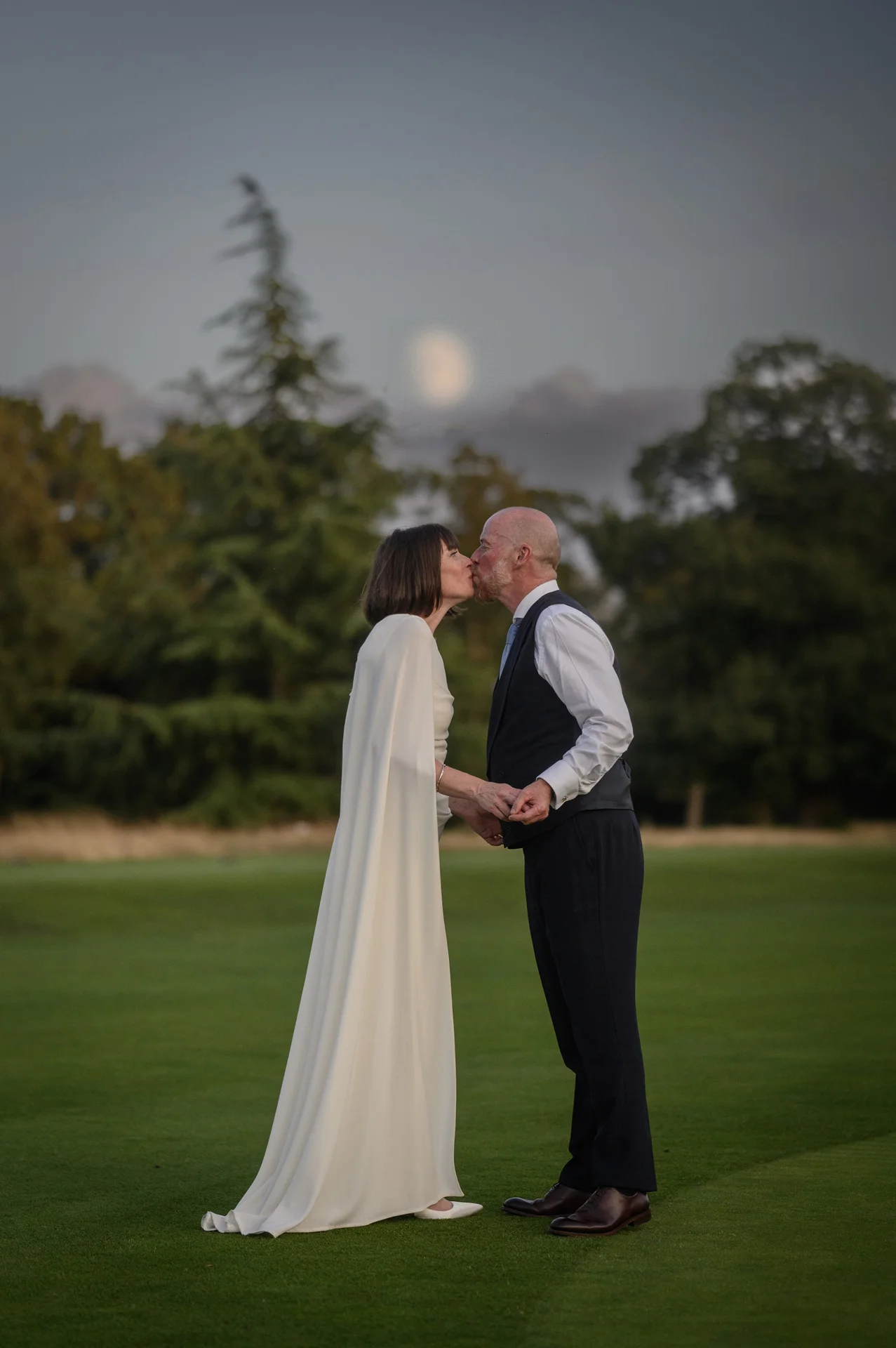 An older couple shares a tender kiss outdoors during dusk, framed against a moody evening sky and distant trees on a manicured lawn.