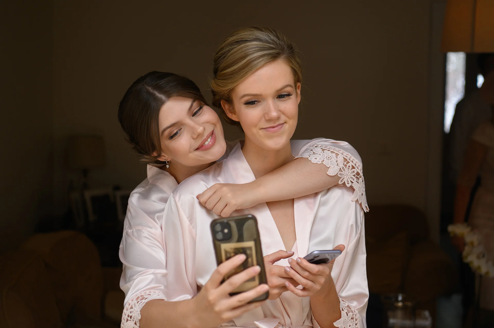 Two women wearing matching satin robes and elegant updos pose together indoors while checking their phones during wedding morning preparations.
