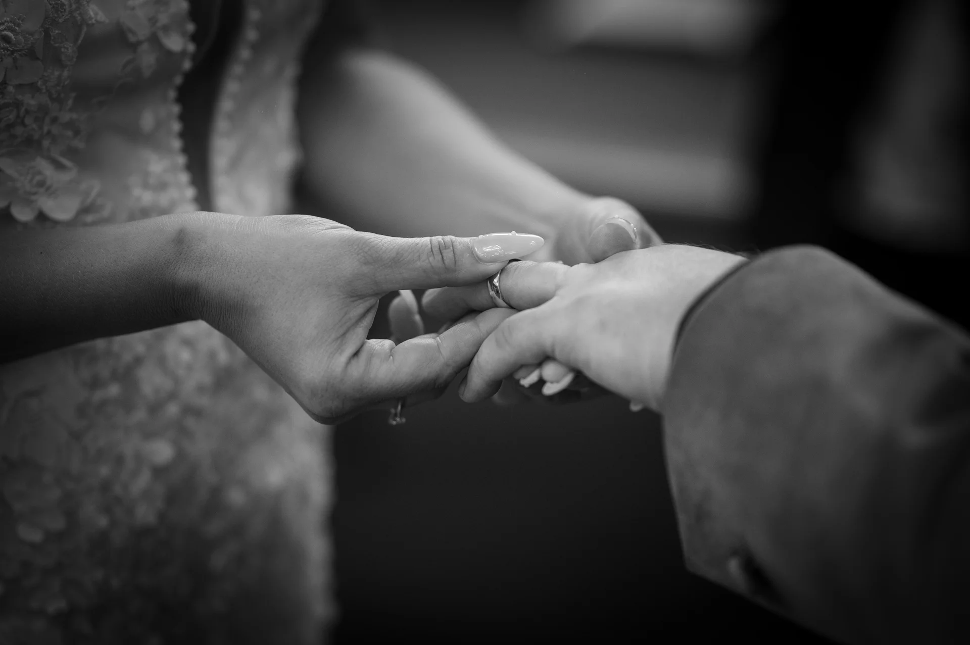 A close up black and white shot captures the tender moment during the ceremony as the bride gently slides a wedding ring onto the groom's finger.