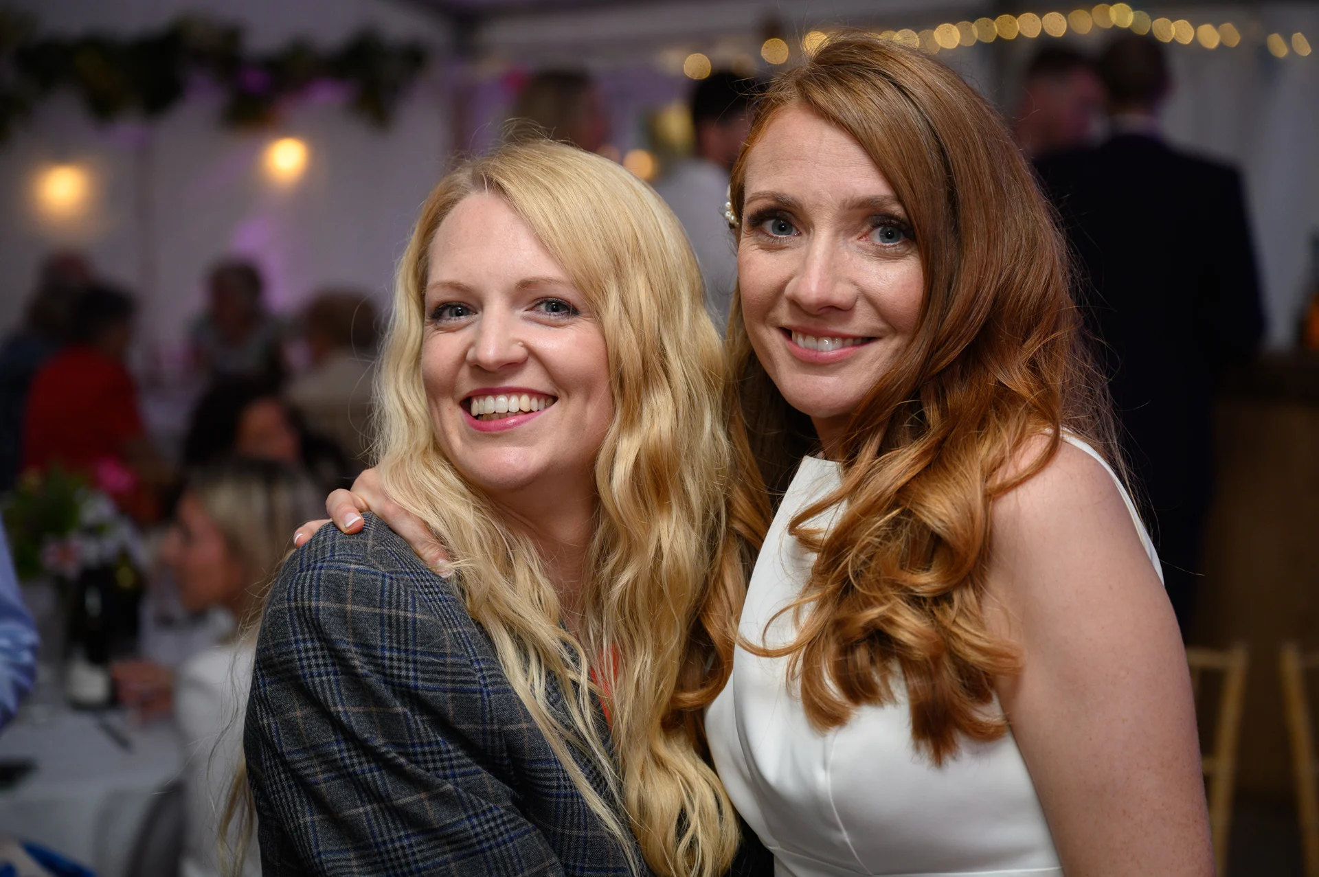 Two smiling brides embrace affectionately during a lively wedding reception, surrounded by guests and softly lit fairy lights.