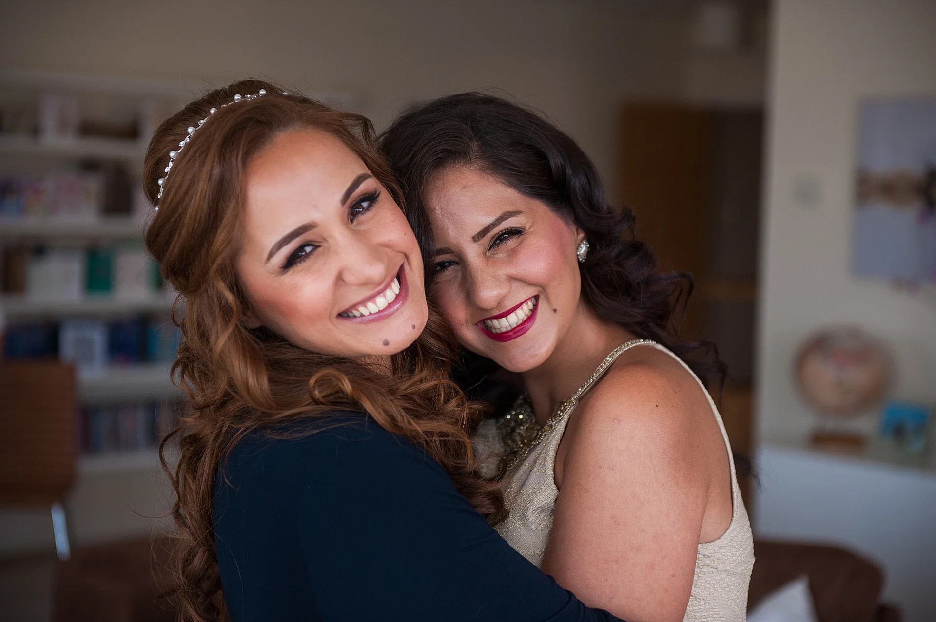 Two smiling brides embrace closely indoors during wedding preparations, both wearing radiant makeup and looking happily towards the camera.