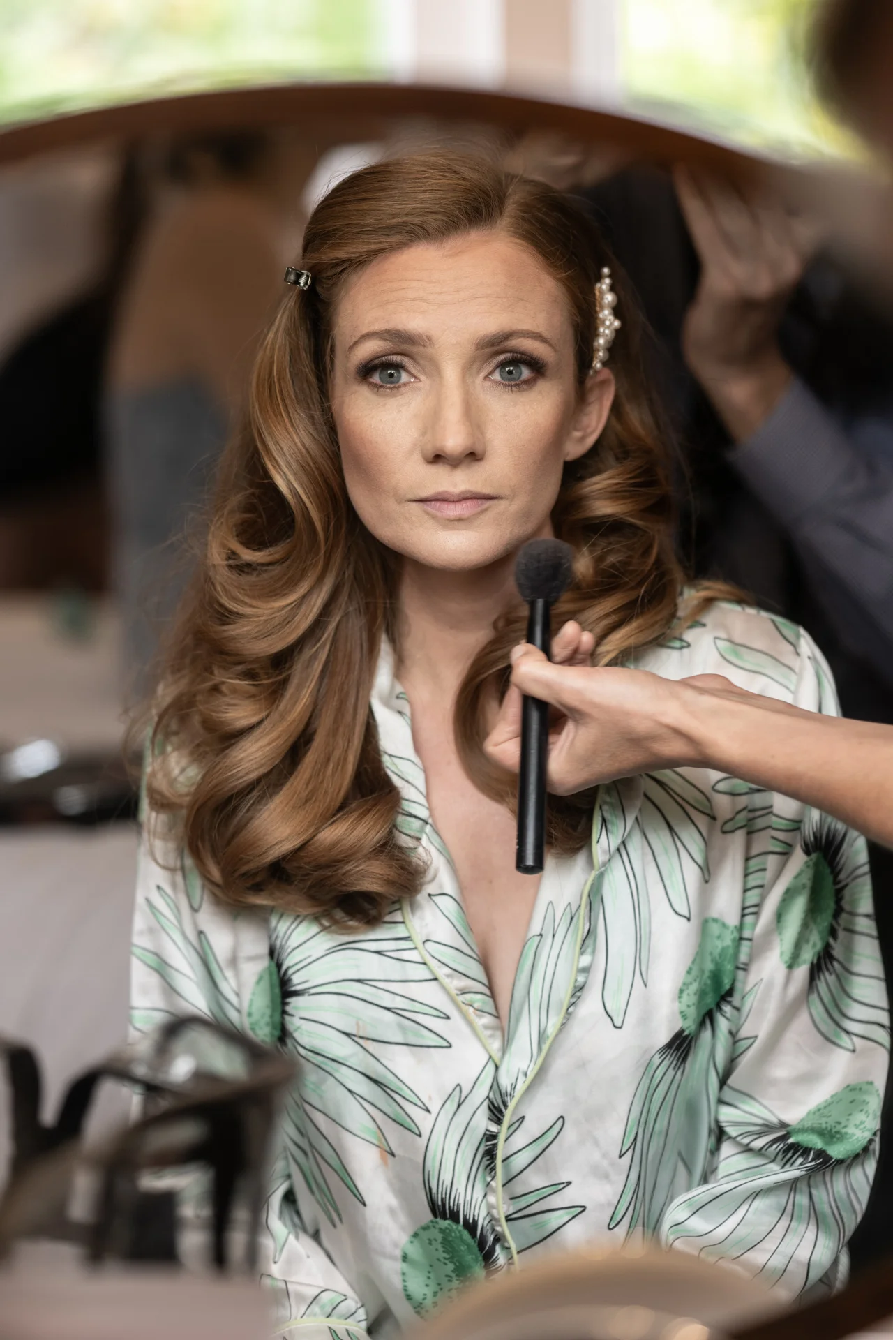 A mature bride sits before a mirror during her wedding morning preparations, wearing a botanical print robe while makeup is applied with a brush.