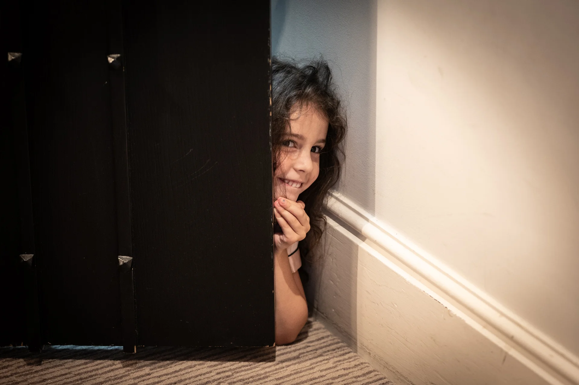 A young flower girl playfully peeks out from behind a dark door, smiling warmly in a candid capture.