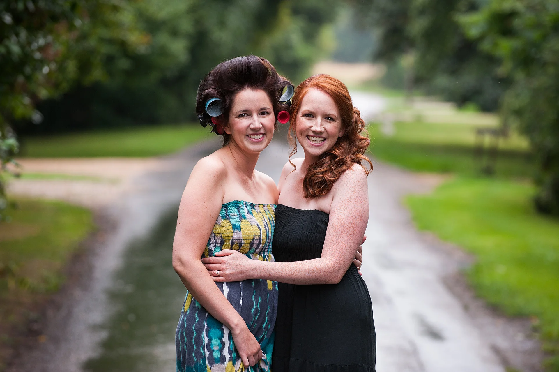 Two smiling women embracing outdoors on a pathway while getting ready for a wedding, with one wearing hair rollers.