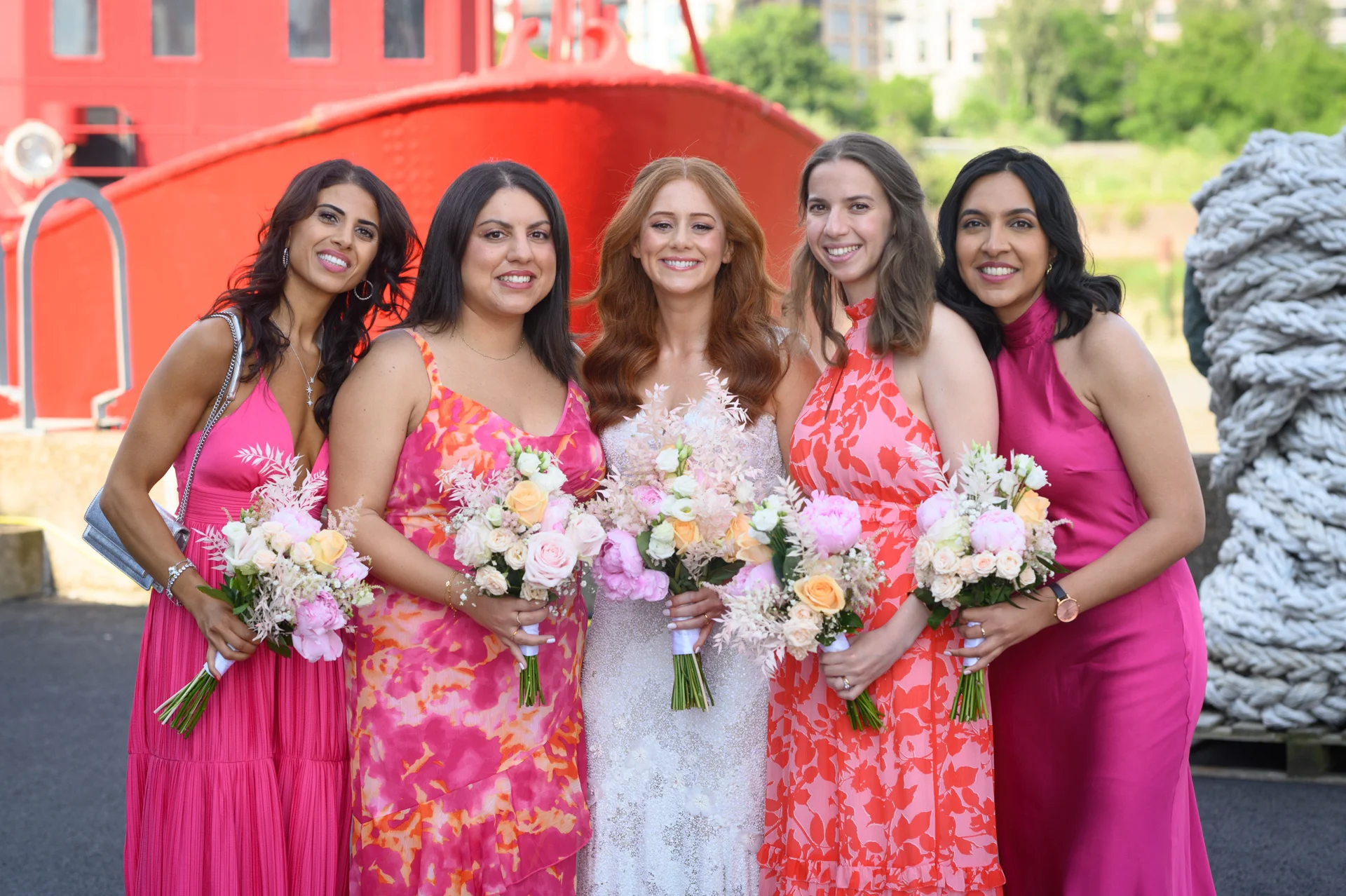 A smiling bride in a textured white gown poses outdoors alongside her four bridesmaids holding matching floral bouquets against a bright red boat backdrop.