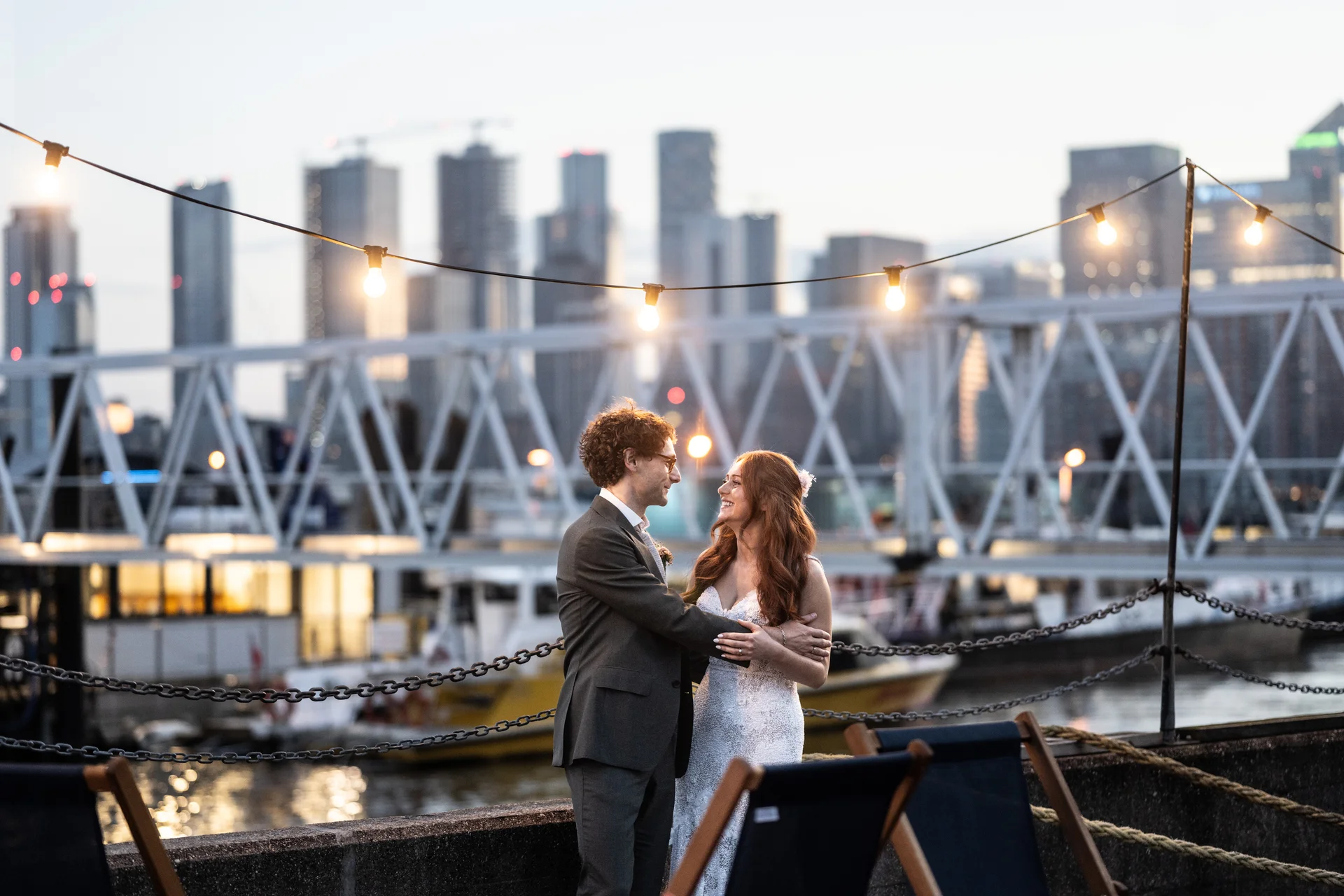 A newlywed couple embraces and smiles lovingly at each other on a waterfront terrace against a glowing city skyline at dusk, framed by overhead festoon lights.