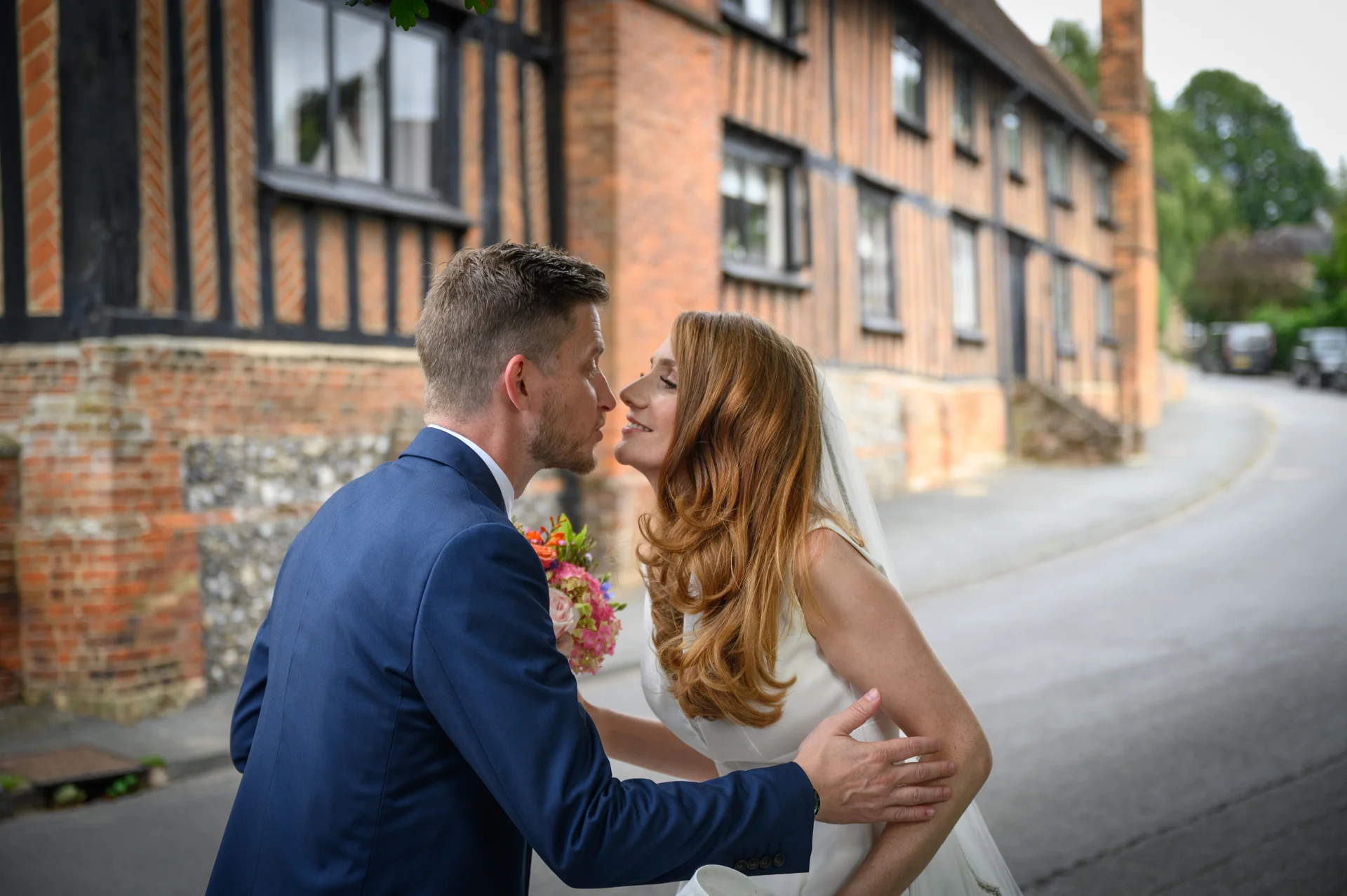 A newlywed couple share a tender moment, standing nose to nose on a village street outside a historic timber-framed building.