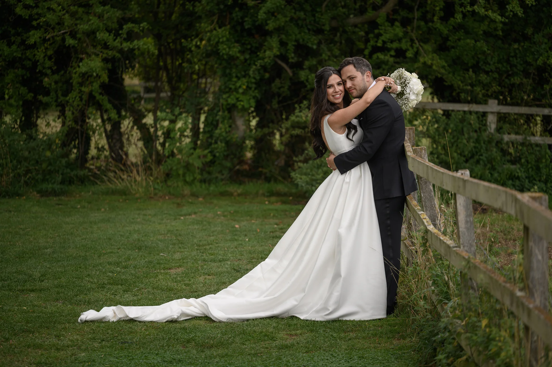 A newlywed couple embraces lovingly next to a rustic wooden fence in a lush green countryside setting, with the bride wearing a classic satin a-line gown and holding a white bouquet.