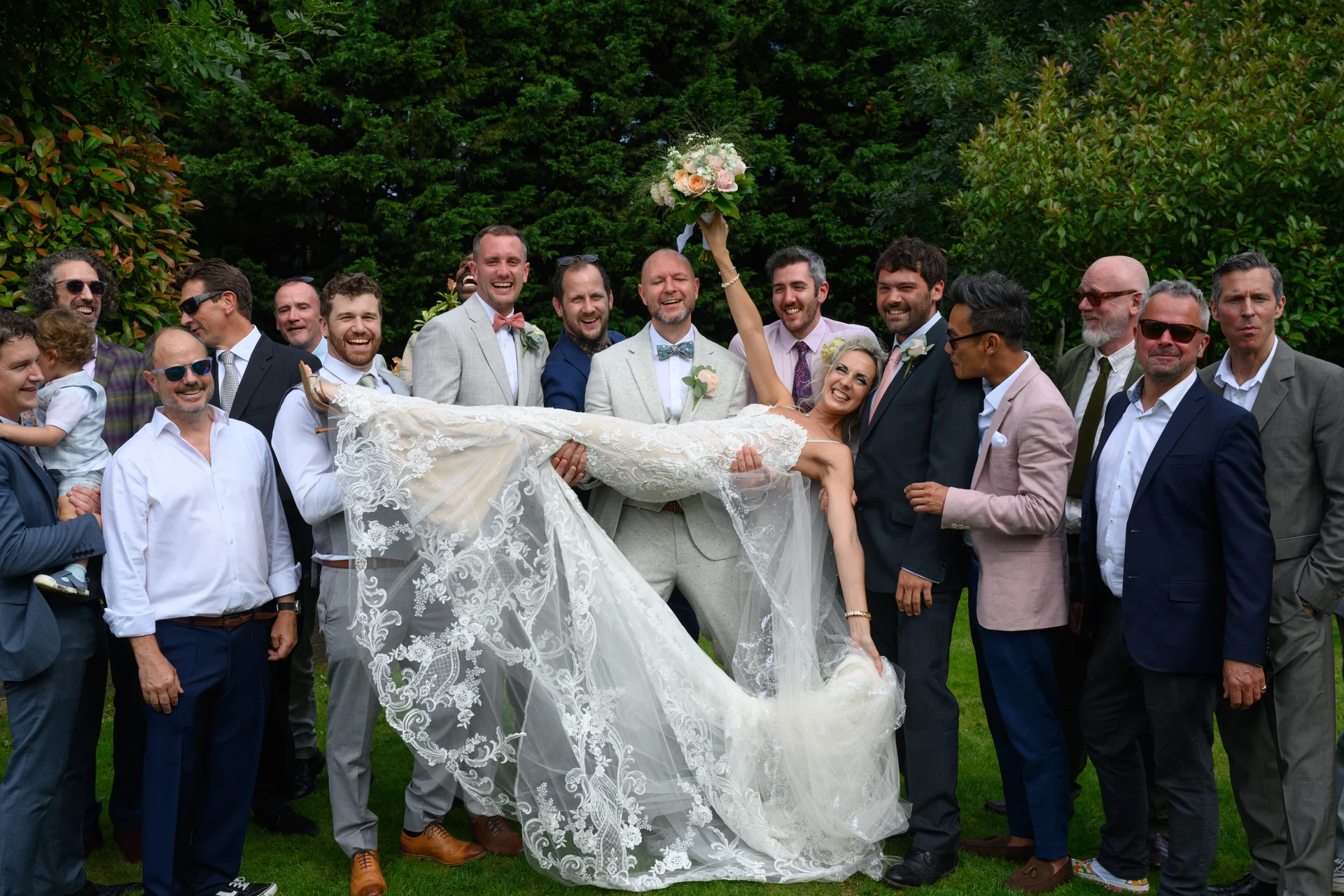 The groom and groomsmen playfully lift the bride horizontally in the air outdoors in a lush garden setting, with the bride holding her bouquet high and smiling joyfully.