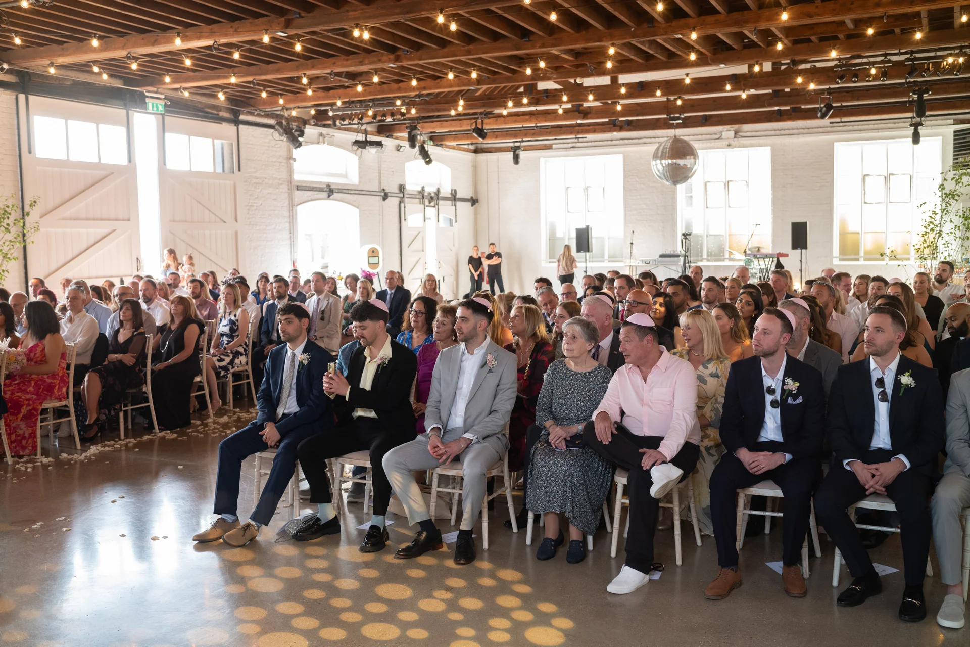 Guests seated in a bright industrial warehouse wedding venue with exposed wooden beams and string lights, awaiting the ceremony.