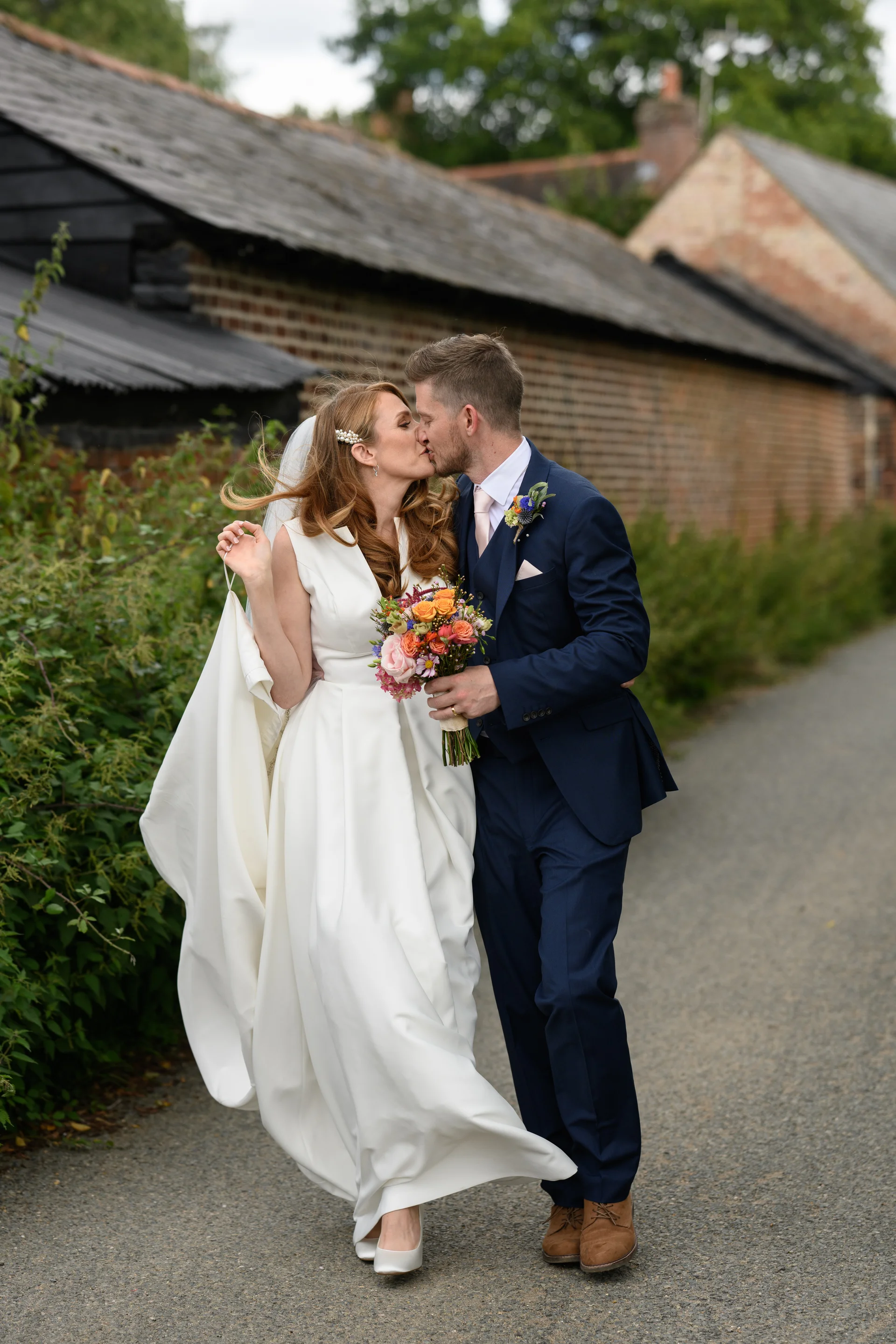 A newlywed couple shares a tender kiss while walking along a gravel path next to rustic brick farm buildings, with the bride holding a colourful bouquet.