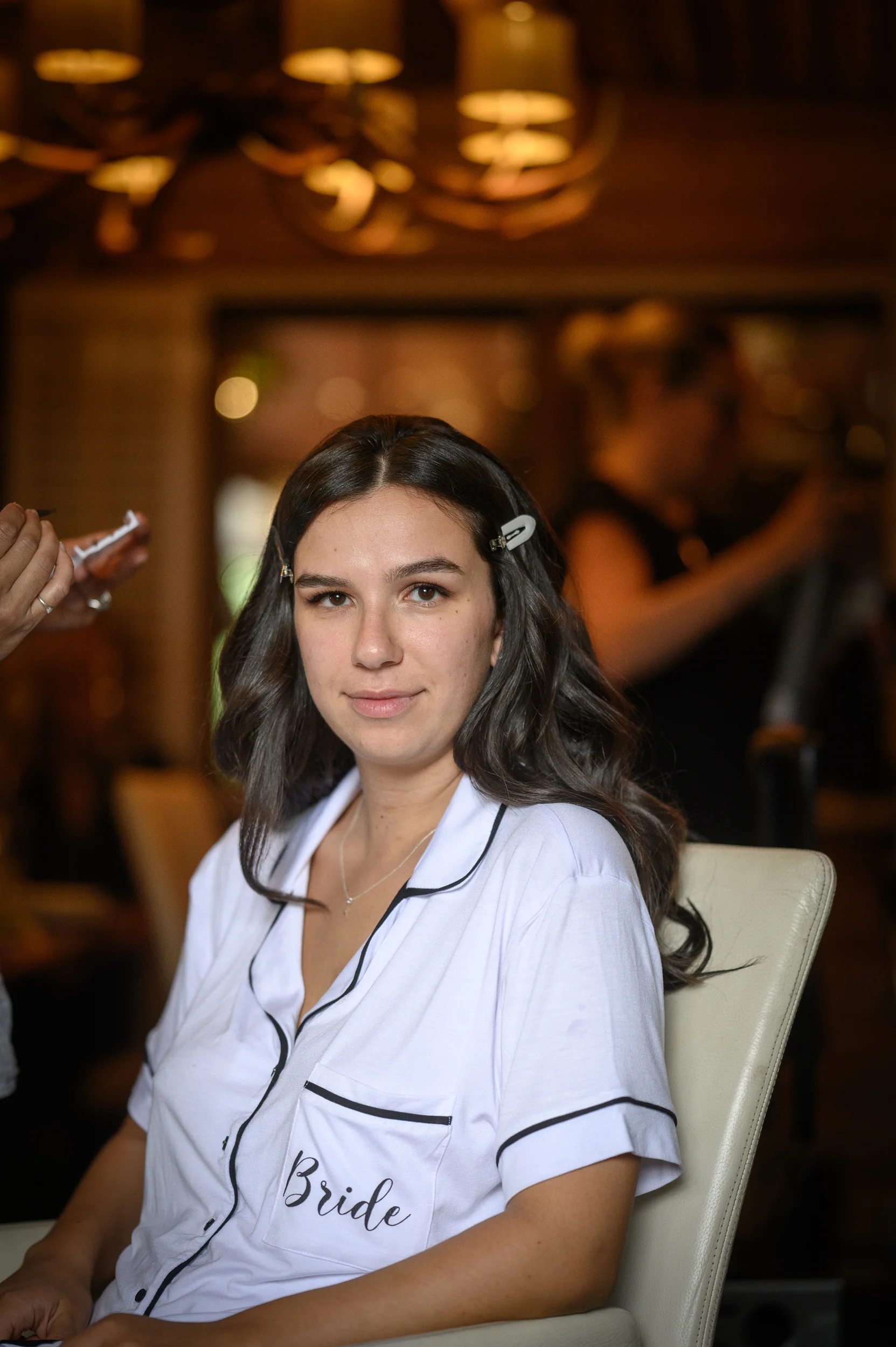A smiling bride with soft dark waves in her hair wears personalised pyjamas while getting her makeup applied during morning bridal preparations.