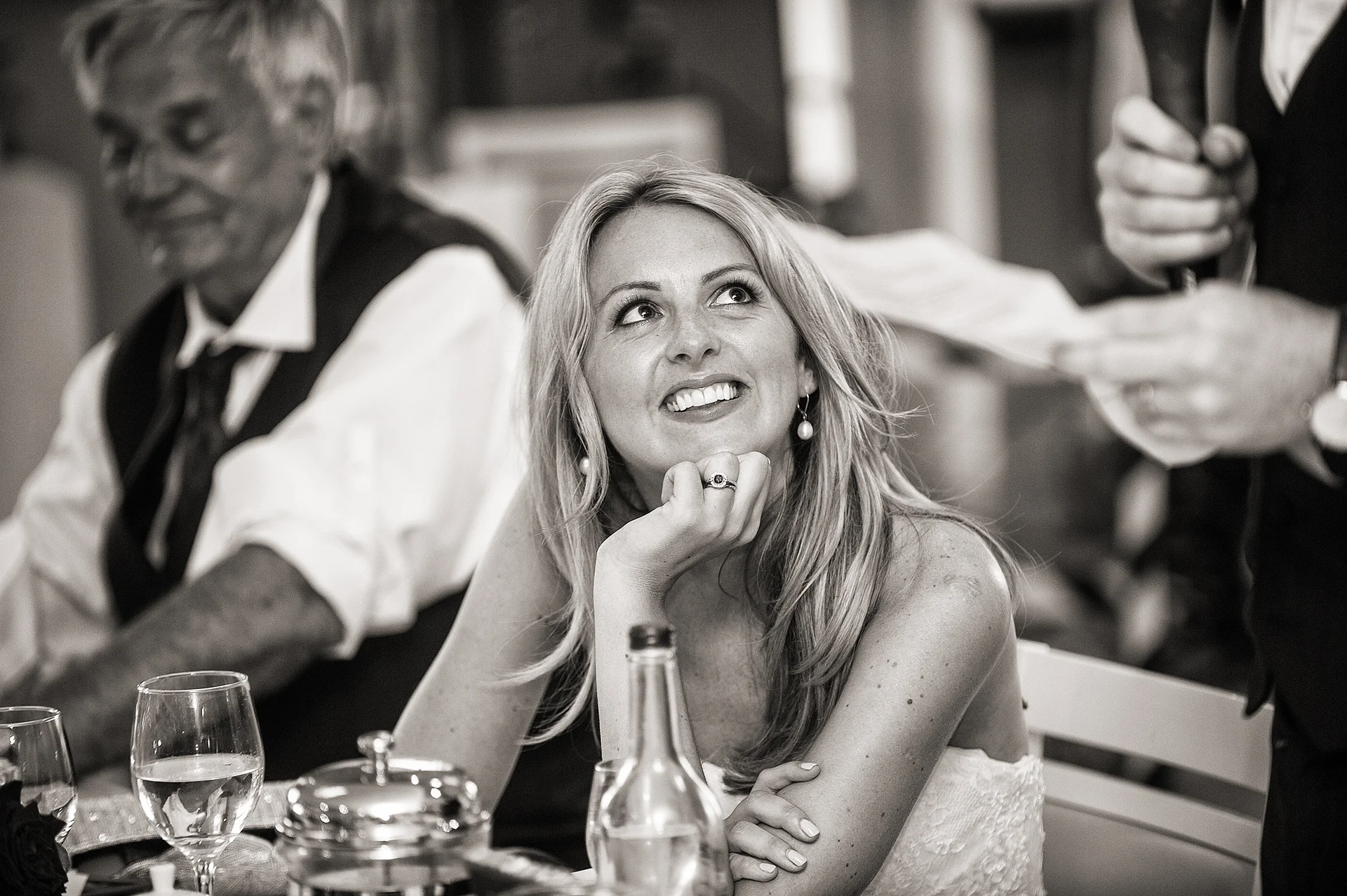 A blonde woman smiling happily with her chin rested on her hand while listening to a wedding speech during the reception.
