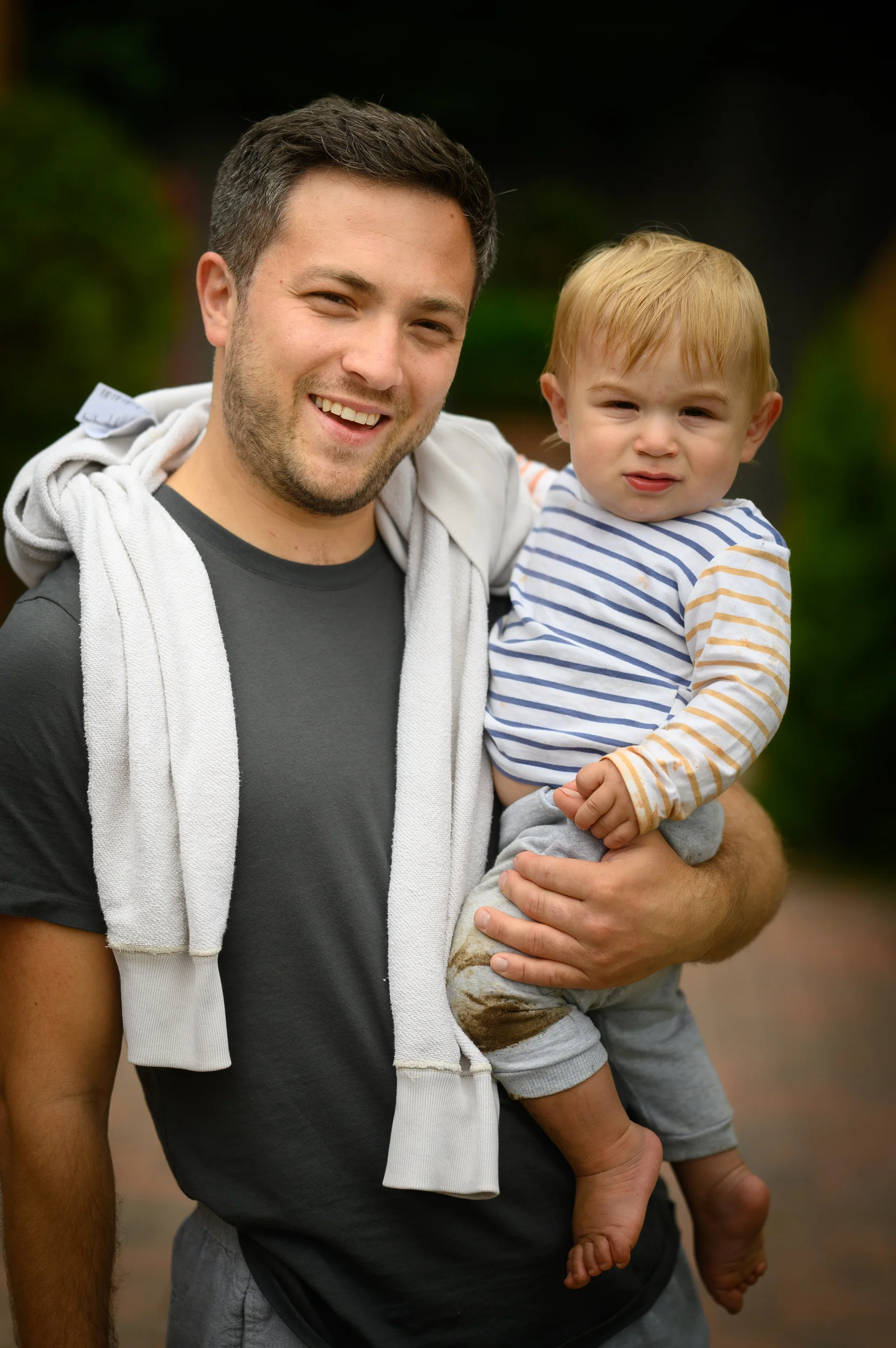A smiling man holding a toddler in a striped top outdoors at a wedding celebration.