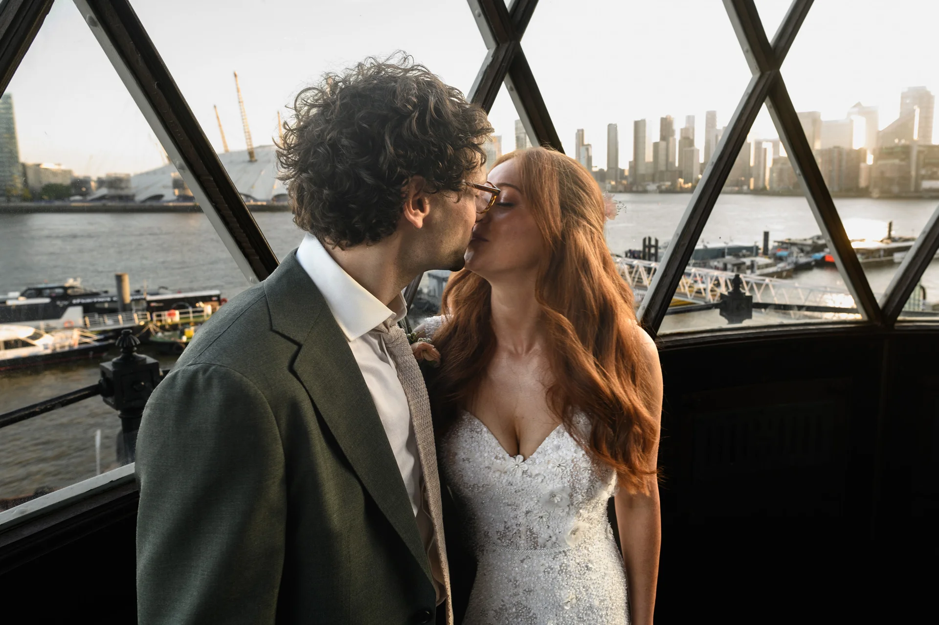 A newlywed couple share a tender kiss inside a glass-paned venue overlooking the London skyline and river during golden hour.