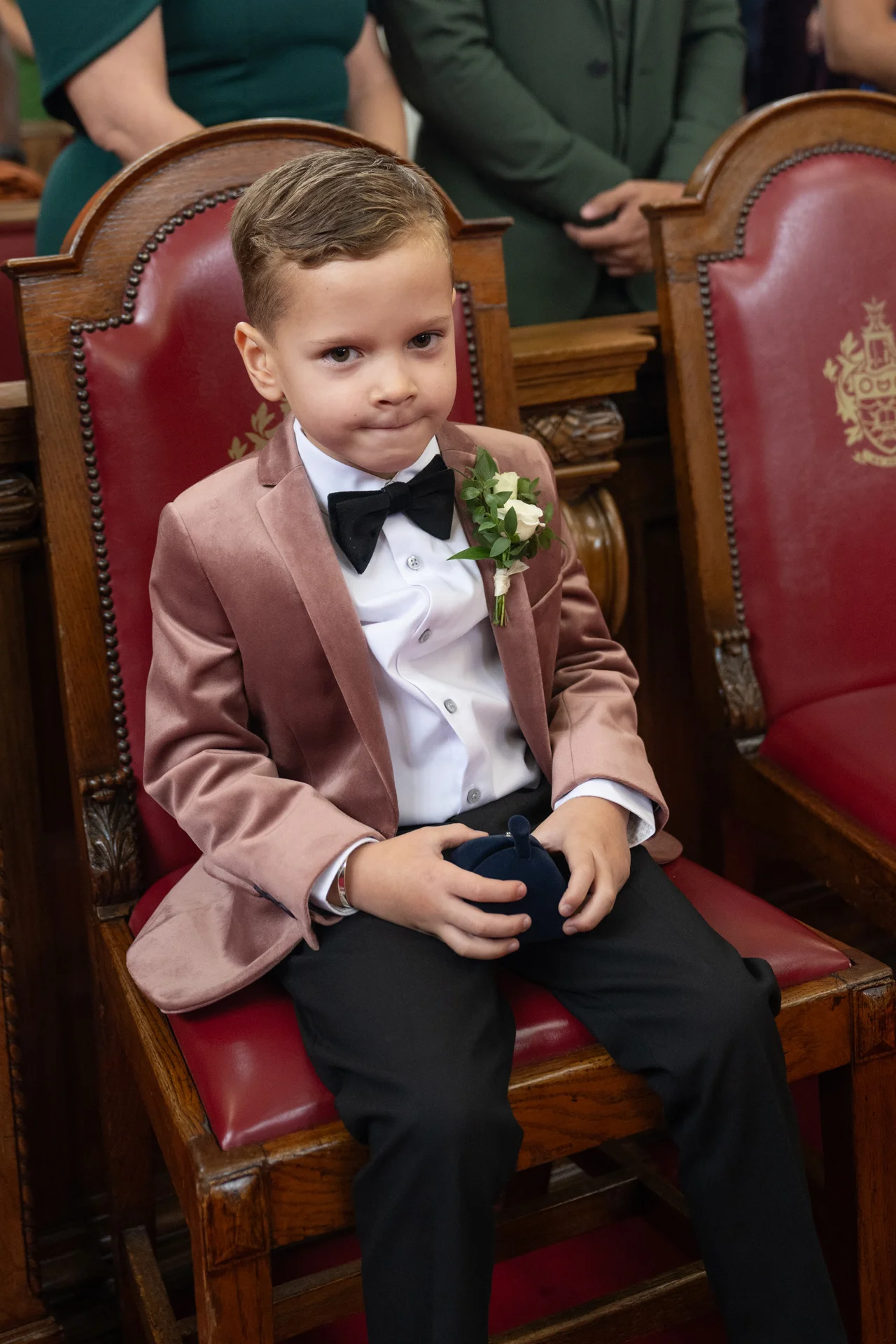 A young page boy dressed in a velvet jacket and bow tie sits on a red ceremony chair holding a ring box.