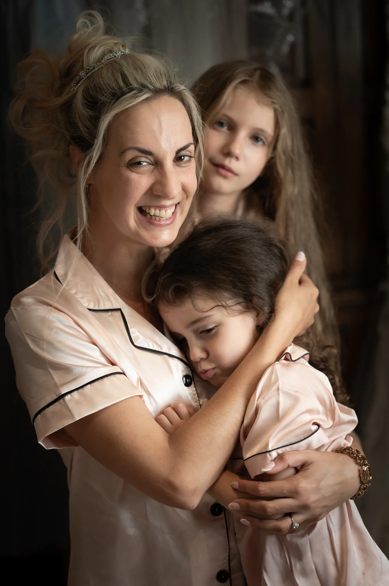 A smiling bride wearing a satin pyjama top embraces a young girl resting on her shoulder during morning wedding preparations, with another child looking on in the background.