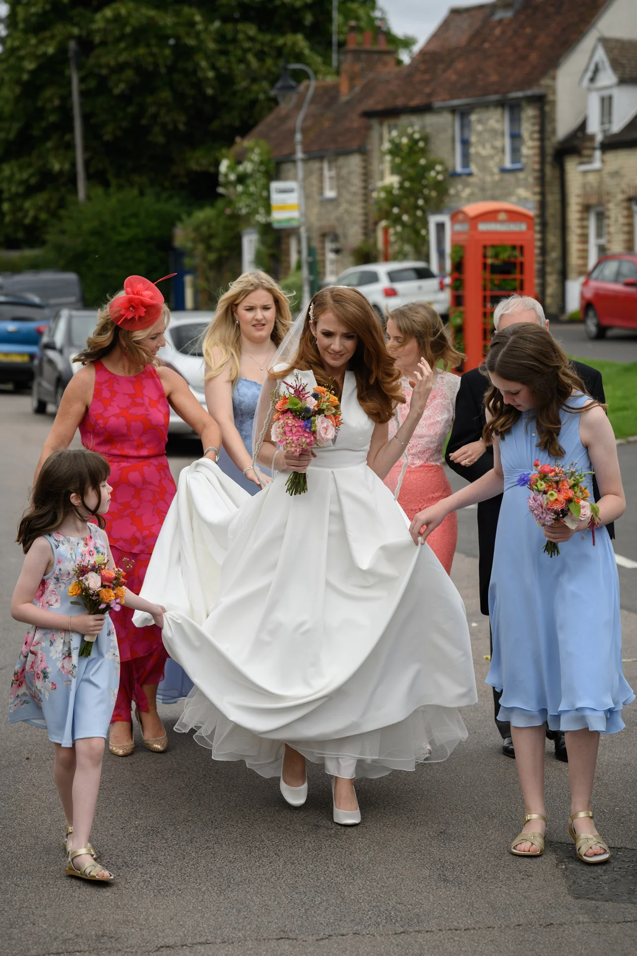 A smiling bride holding a colourful bouquet walks down a village street surrounded by bridesmaids and a flower girl in blue and pink dresses, near a traditional red telephone box.