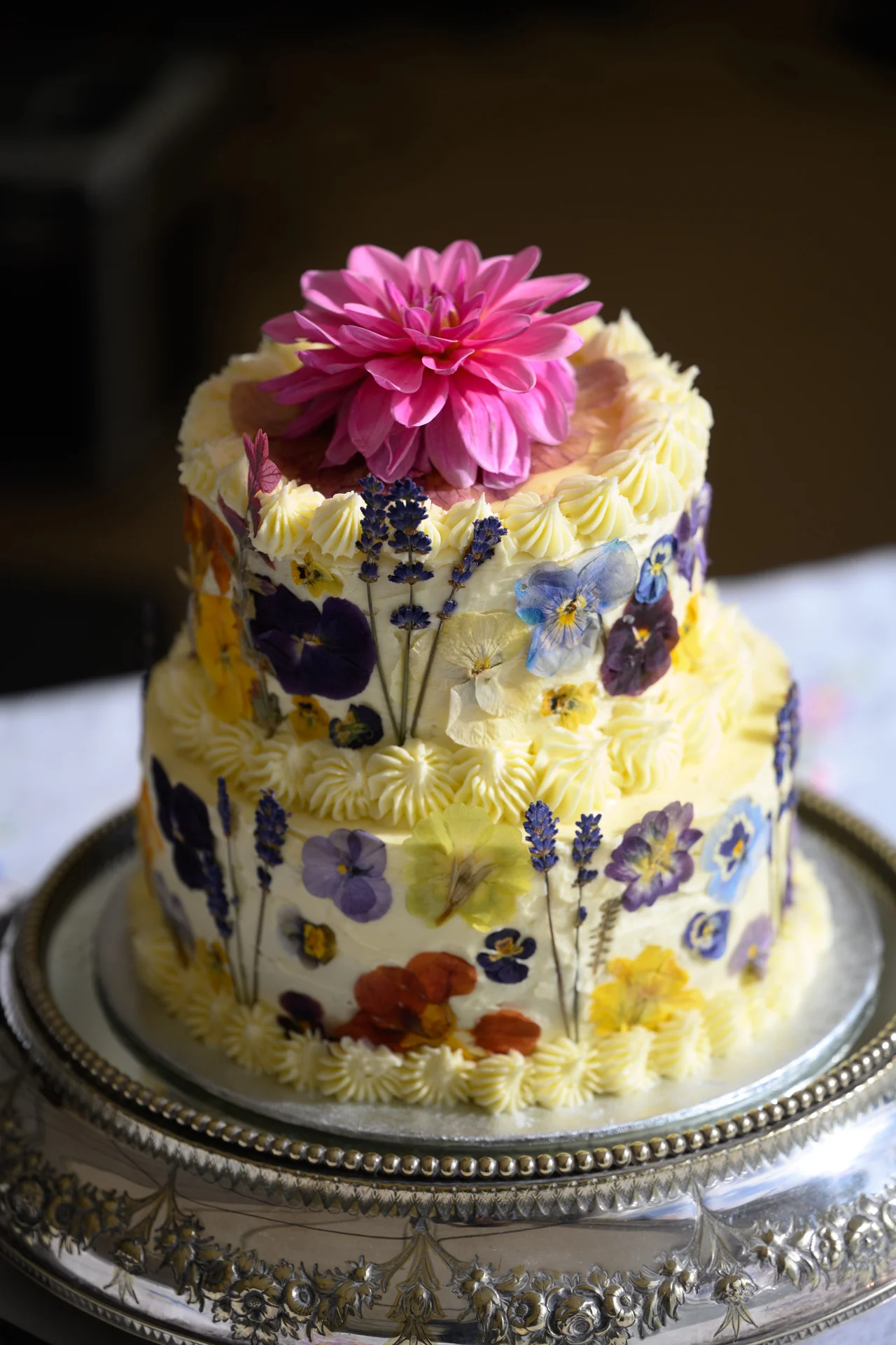 A stunning two-tier buttercream wedding cake decorated with pressed flowers and topped with a vibrant pink dahlia sits on a decorative silver cake stand.
