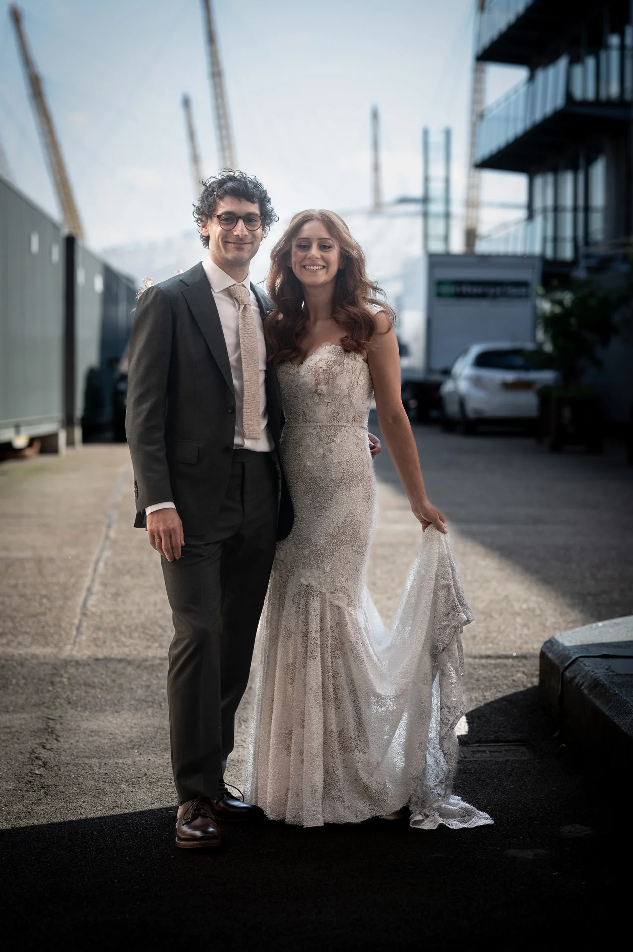 A newlywed couple poses together in an urban industrial setting with the bride holding her textured lace mermaid wedding dress.
