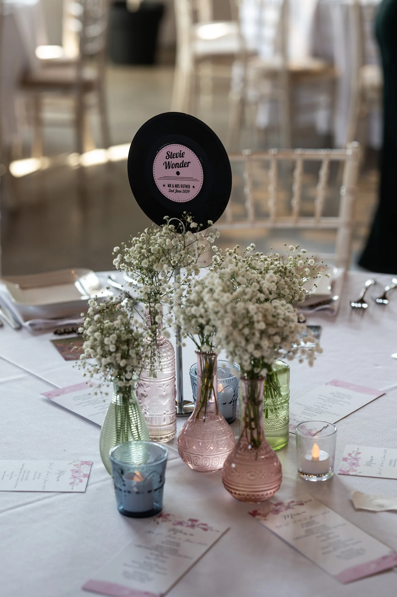 A wedding reception table centerpiece featuring multiple pink and green bud vases filled with baby's breath, surrounded by votive candles, menu cards, and a vinyl record table name sign.