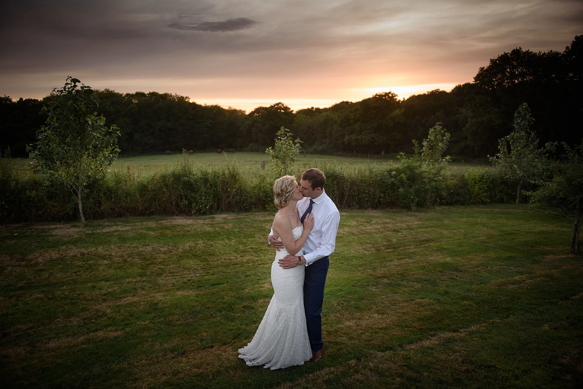 A newlywed couple shares a tender kiss standing on a grassy lawn during a dramatic sunset against a scenic countryside backdrop.