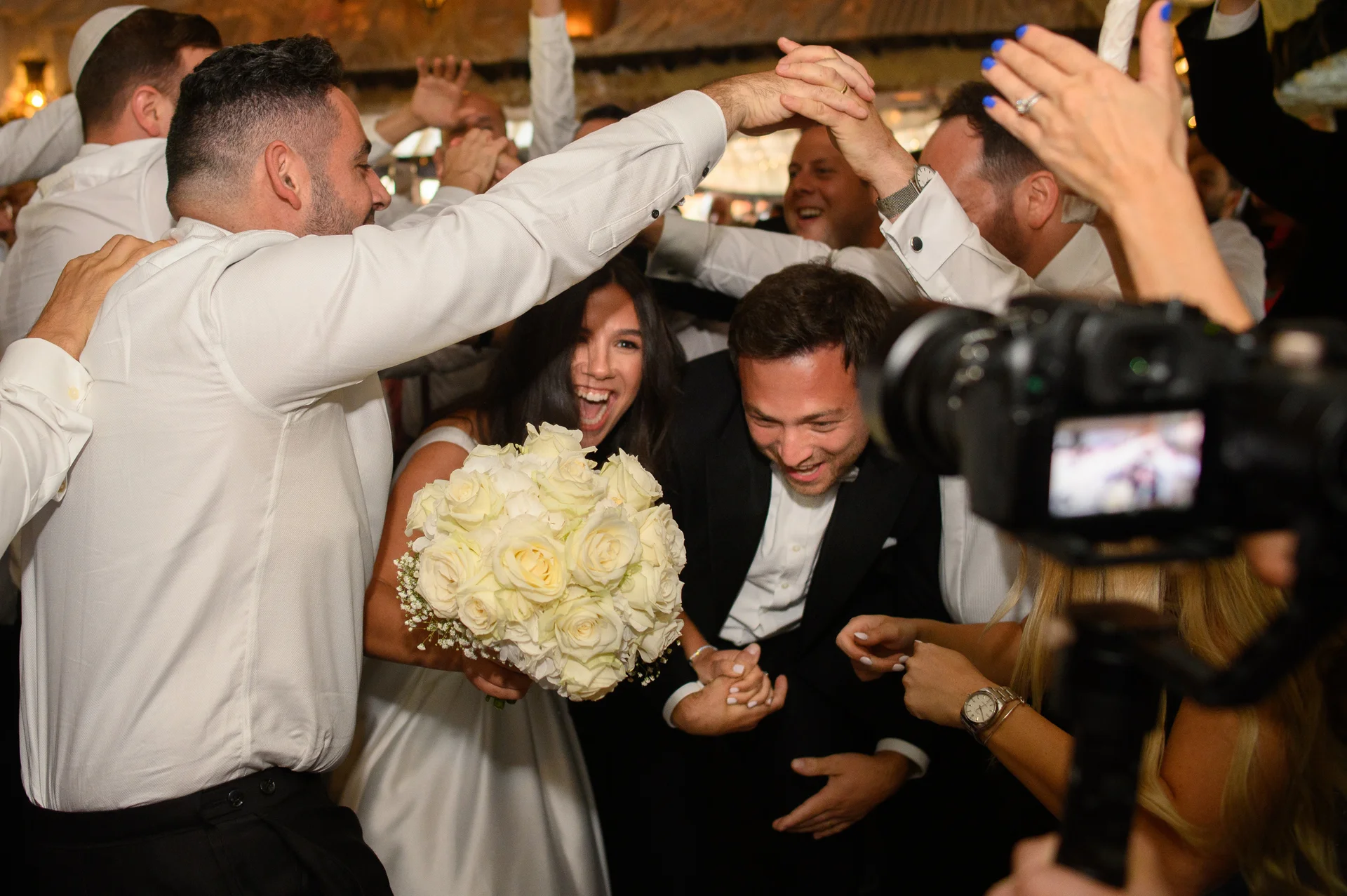 A joyful bride and groom are surrounded by cheering wedding guests participating in traditional circle dancing with the groom laughing heartily.
