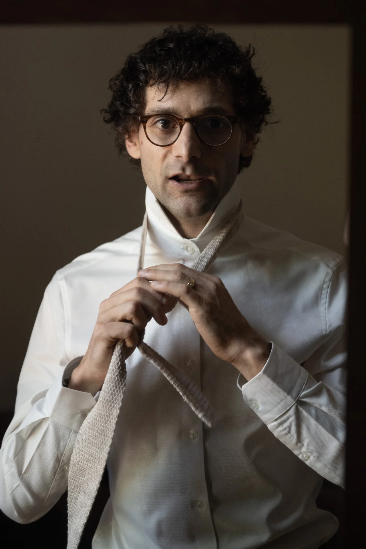 A groom with curly hair and glasses stands in front of a mirror while tying his textured neutral-toned tie during morning preparations.