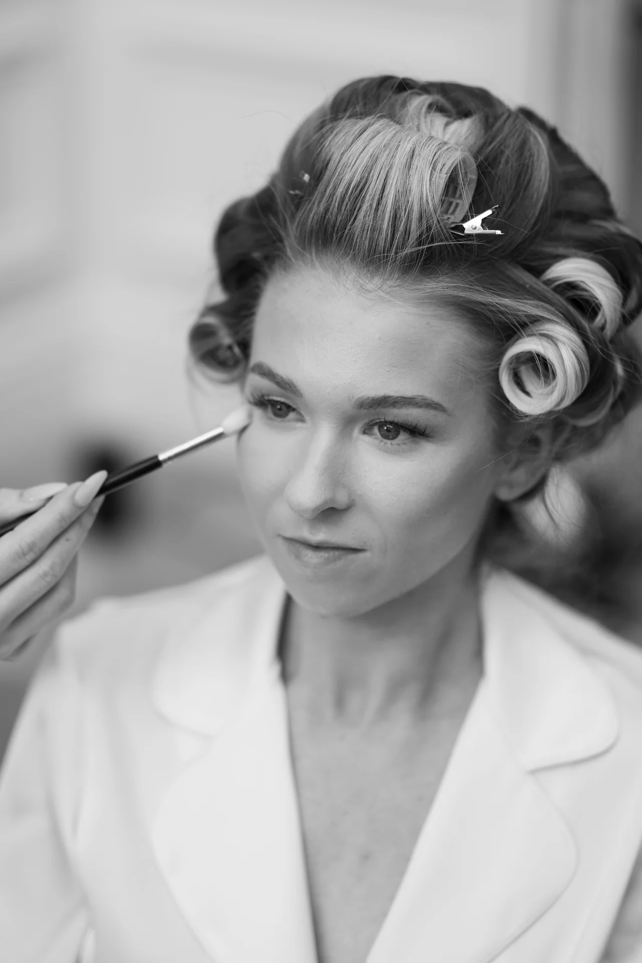 A close-up black and white shot of a bride with hair rollers having her makeup applied with a brush during morning preparations.