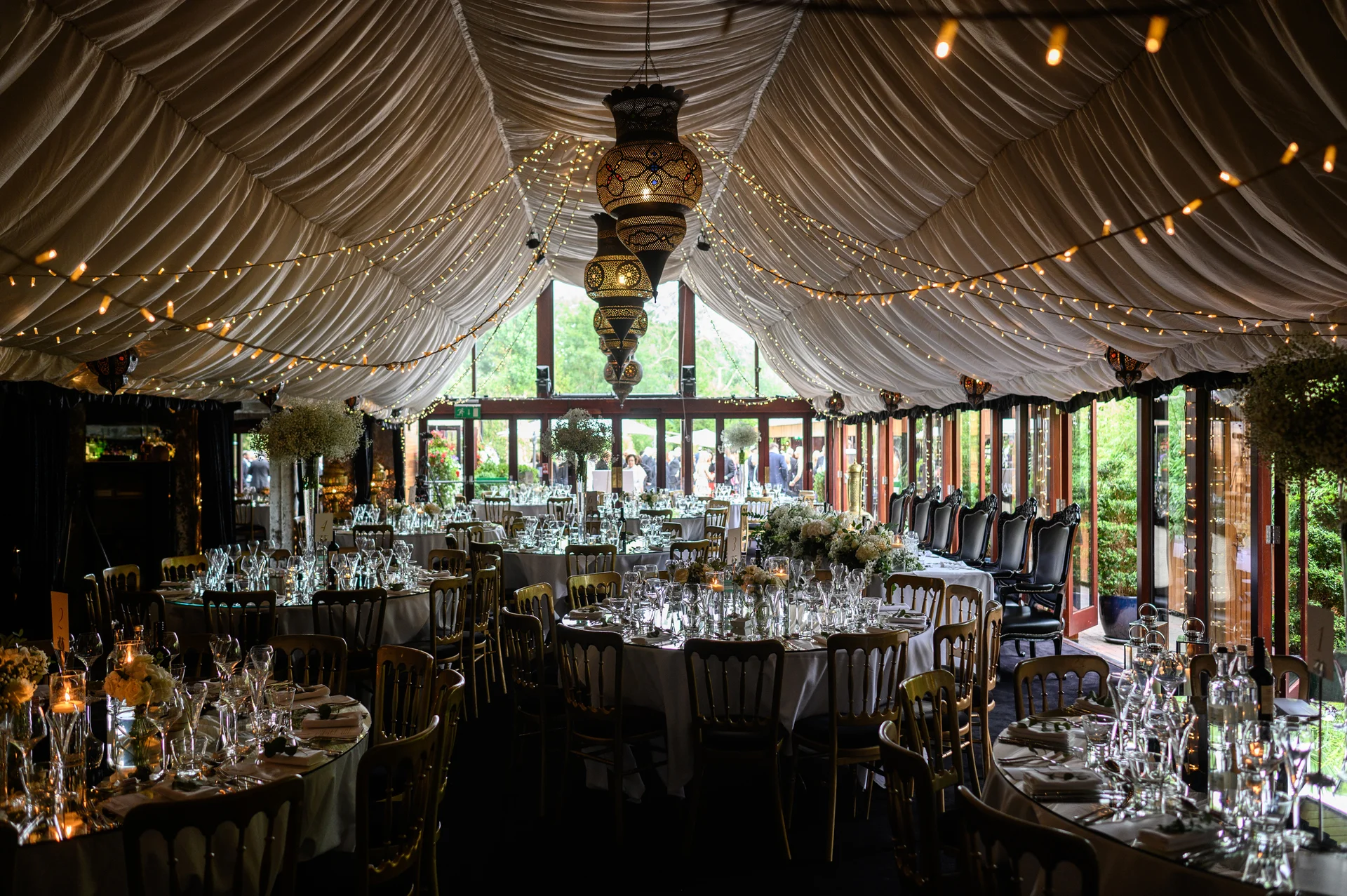 An elegant marquee wedding reception venue features draped white ceiling fabric, sparkling fairy light canopies, hanging lanterns, and beautifully set round dining tables.