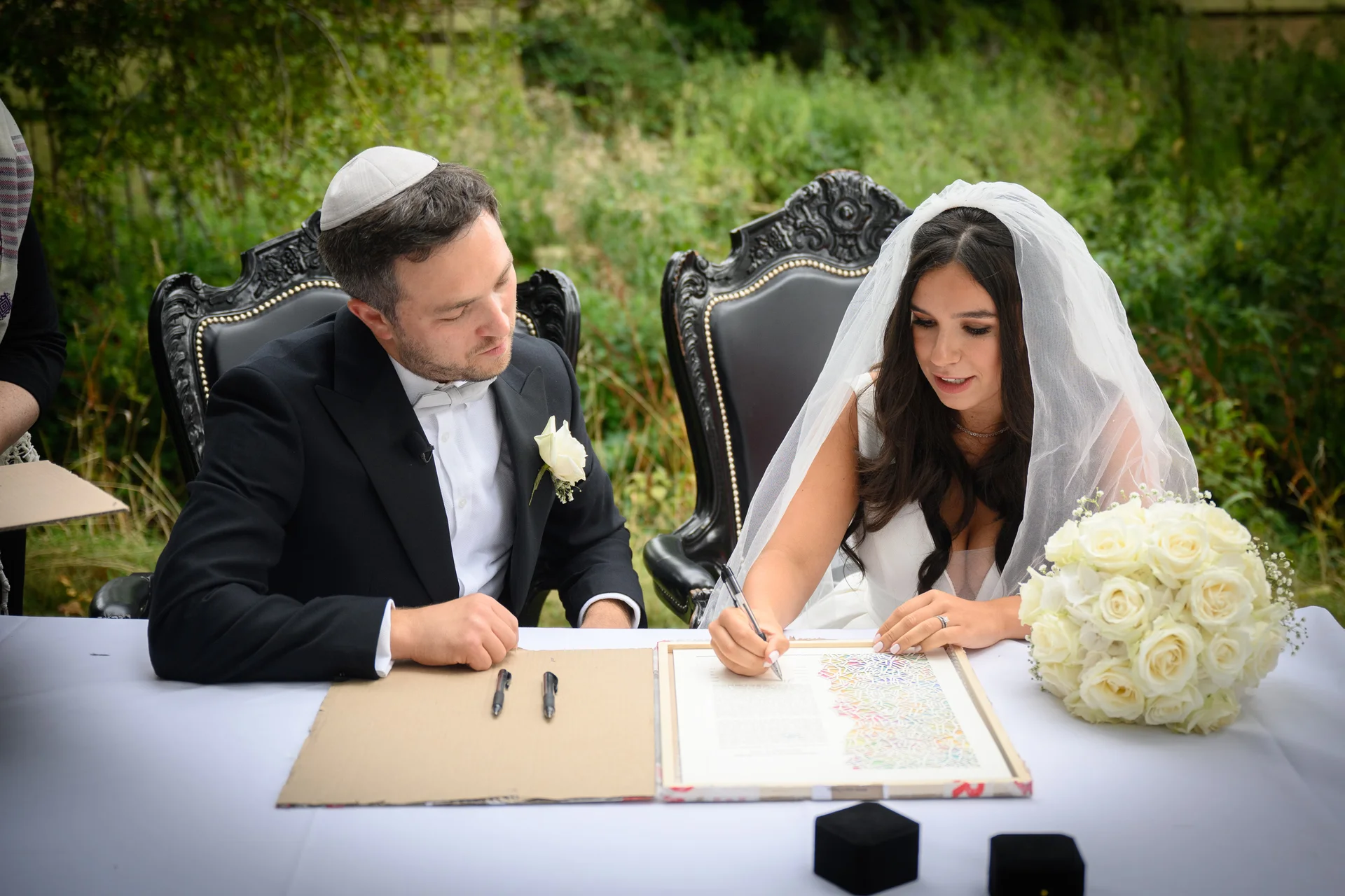 A bride in a white veil and a groom wearing a kippah sit side by side at a white-clothed table outdoors, signing their ketubah next to a bouquet of white roses.