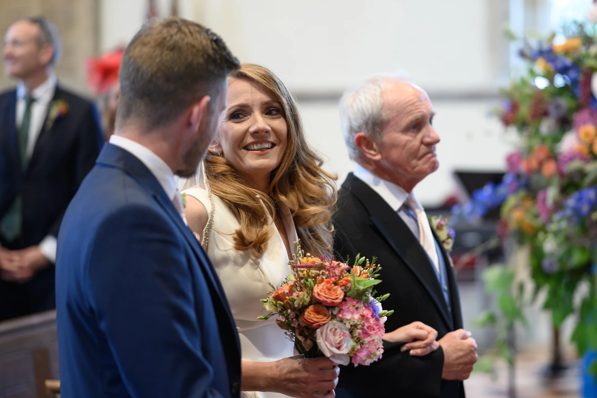 A joyful bride holding a colourful hand-tied bouquet shares a warm glance with her groom during the wedding ceremony alongside her father.