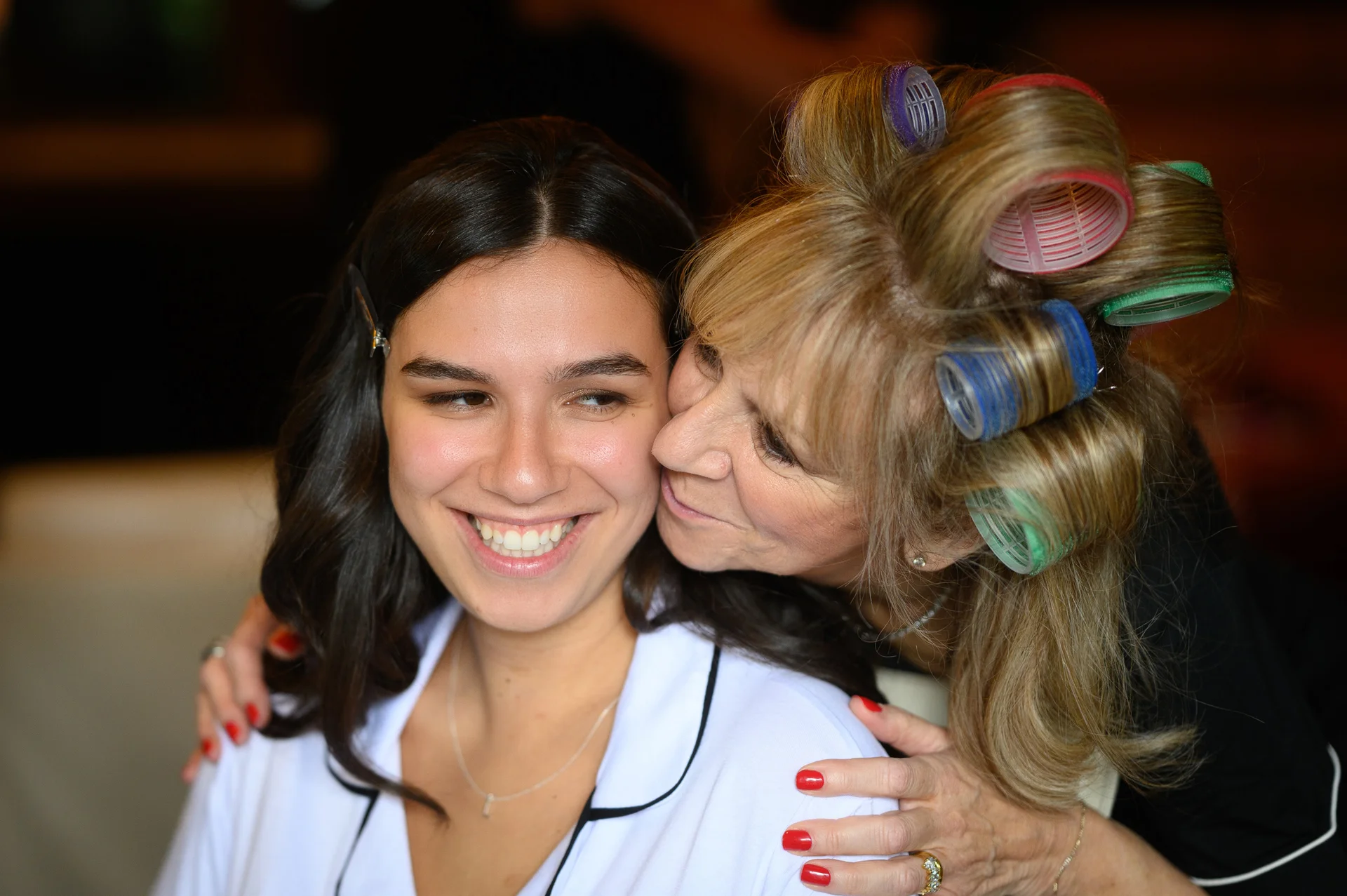 An older woman with hair rollers in her hair affectionately embraces and kisses the cheek of a smiling young woman wearing a white robe during bridal morning preparations.