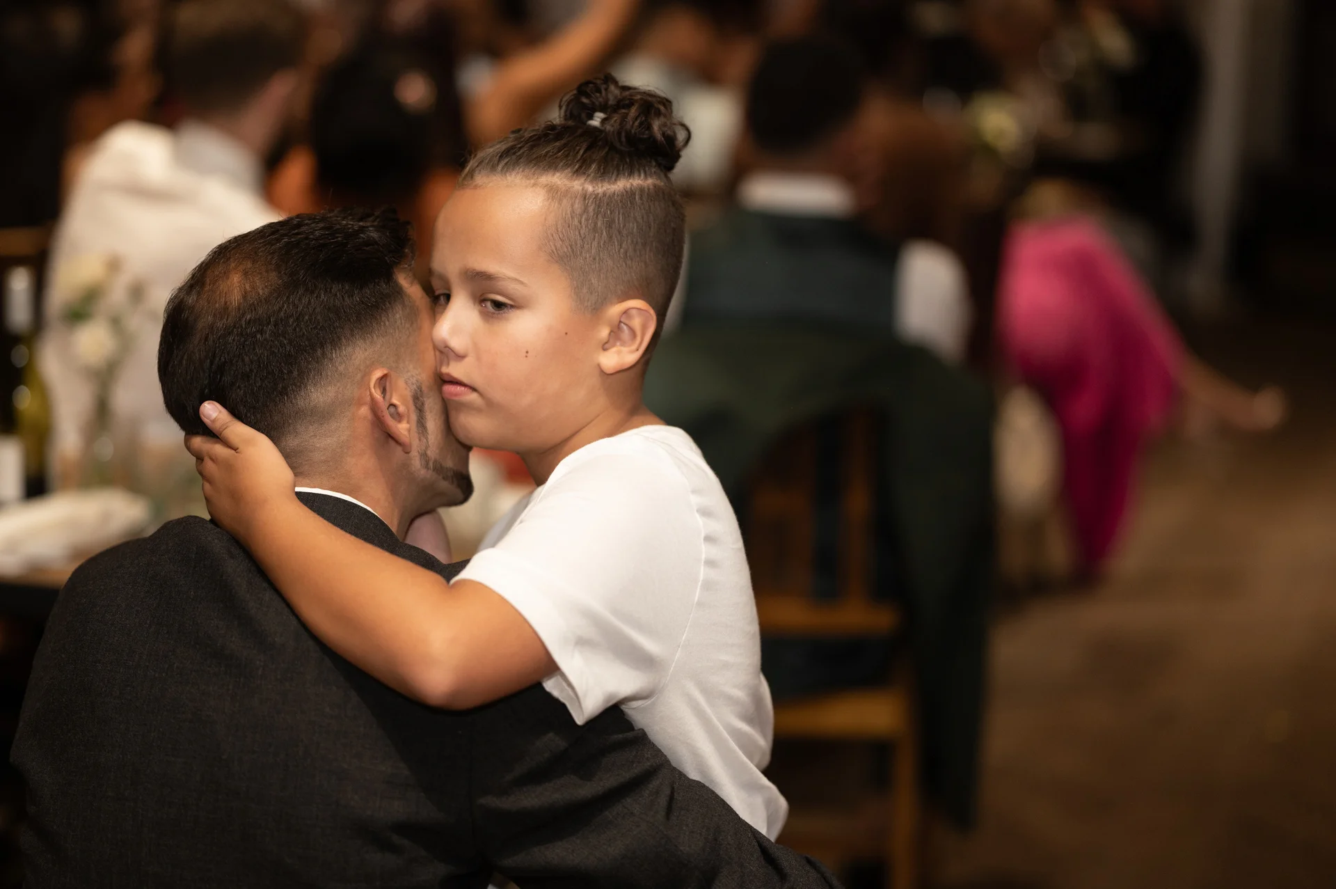A young boy embraces an adult in a dark suit tightly during a wedding reception, sharing a tender and emotional moment amidst blurred guests.