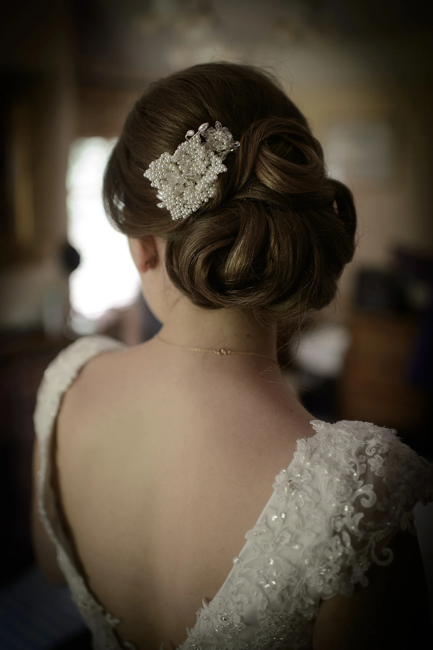 A back view of a bride showcasing her elegant low chignon updo adorned with an intricate pearl hair comb and a detailed lace gown with an open back.